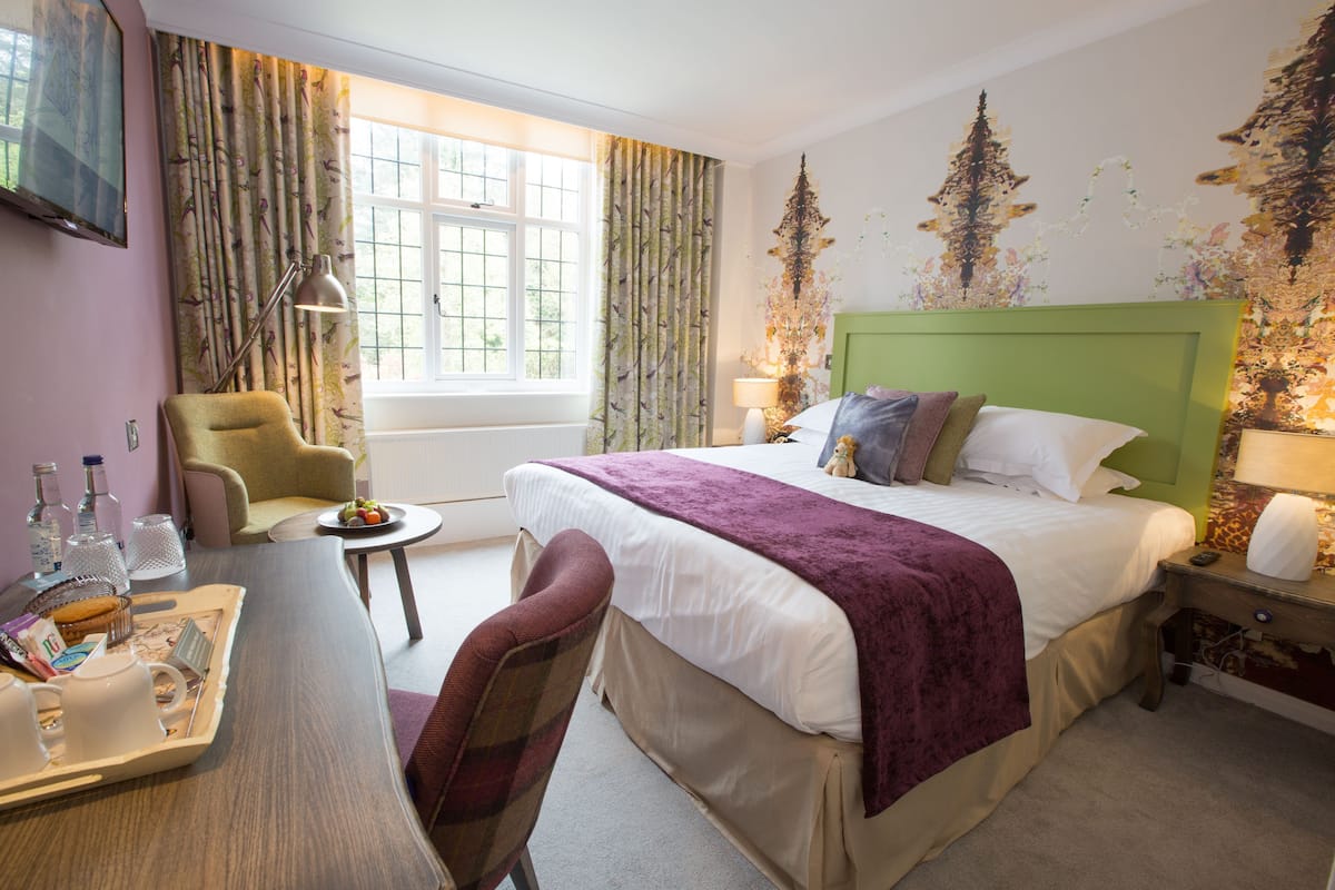 A hotel room with a bed, a desk with a tray of breakfast items, a chair, a lamp, and a window with curtains.