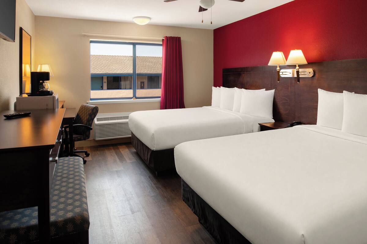 Habitación de hotel con dos camas, un escritorio, una silla, una ventana con cortinas y una lámpara en la pared.