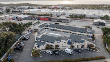 An aerial view of a commercial building with a parking lot, surrounded by greenery and a body of water in the background.