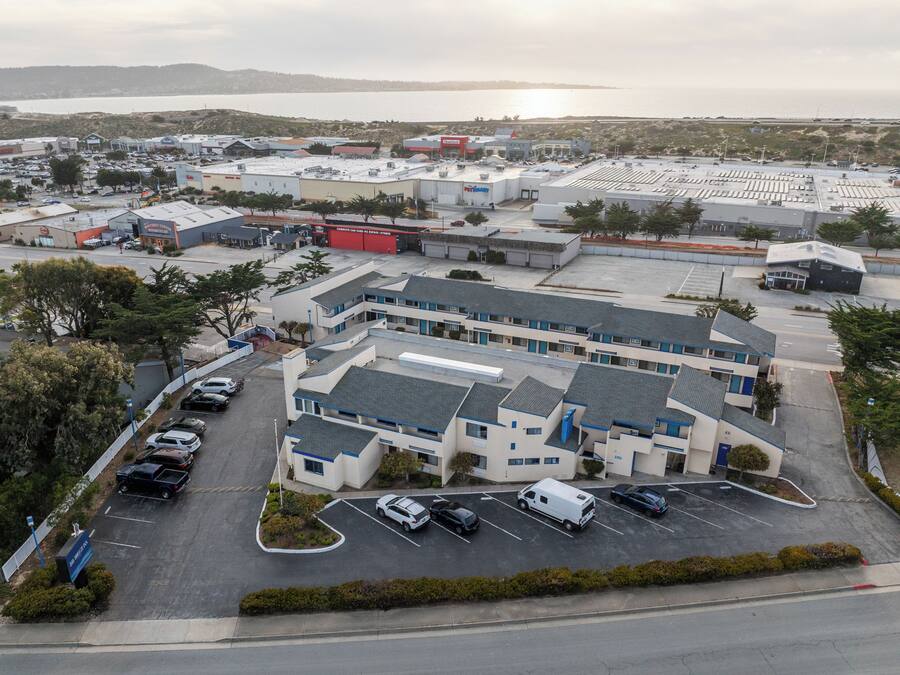 An aerial view of a commercial building with a parking lot, surrounded by greenery and a body of water in the background.