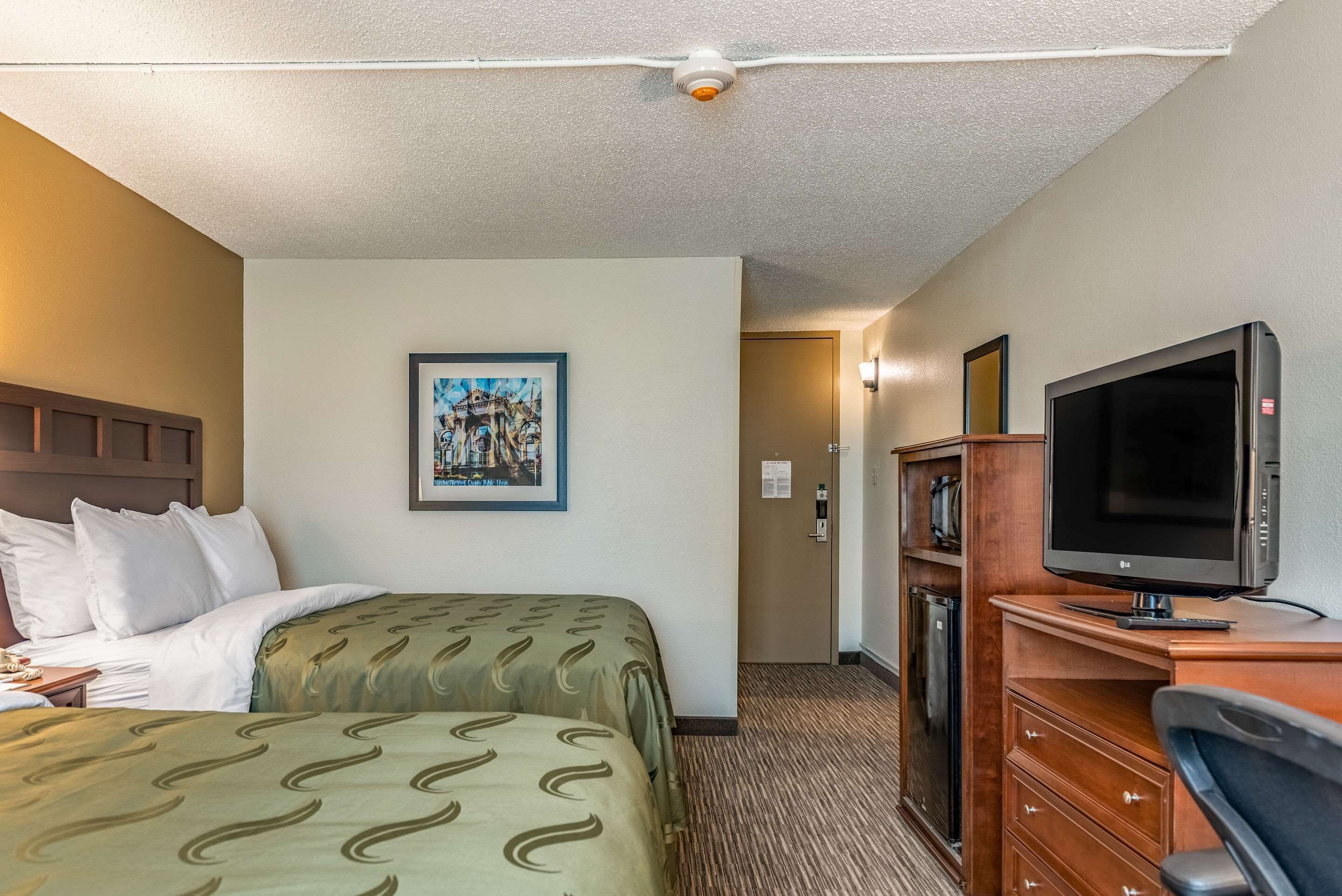 A hotel room with a bed, a desk with a television, a framed picture on the wall, and a door leading to the hallway.