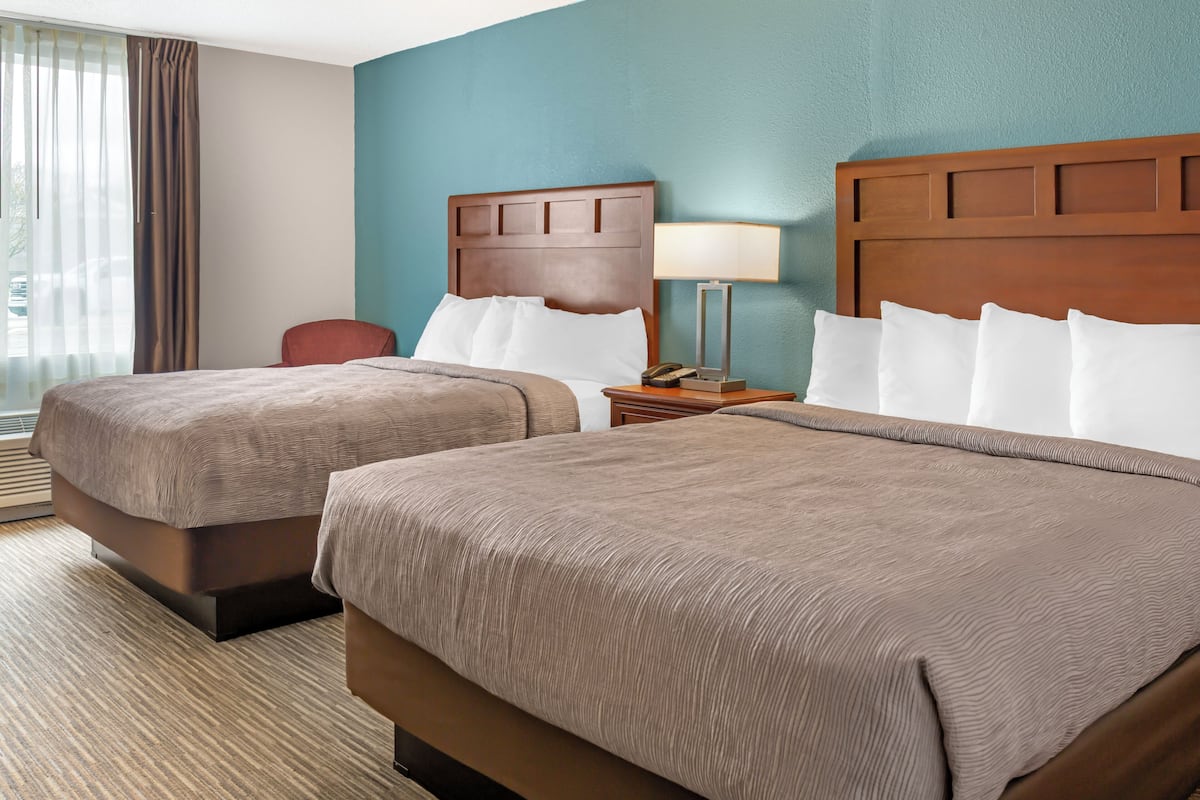 A hotel room with two beds, a nightstand with a lamp, a chair, and a view of the outdoors.