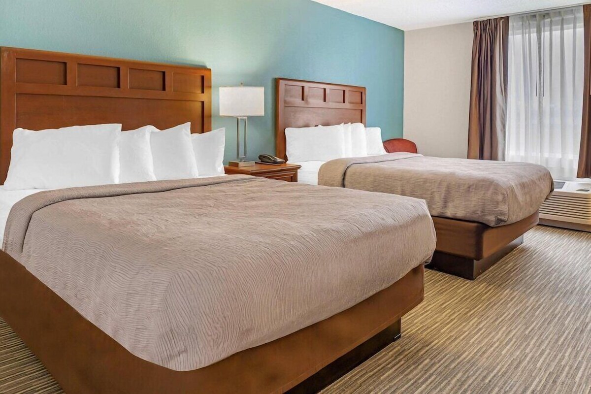 A hotel room with two beds, wooden headboards, a nightstand with a lamp, and a window with curtains.
