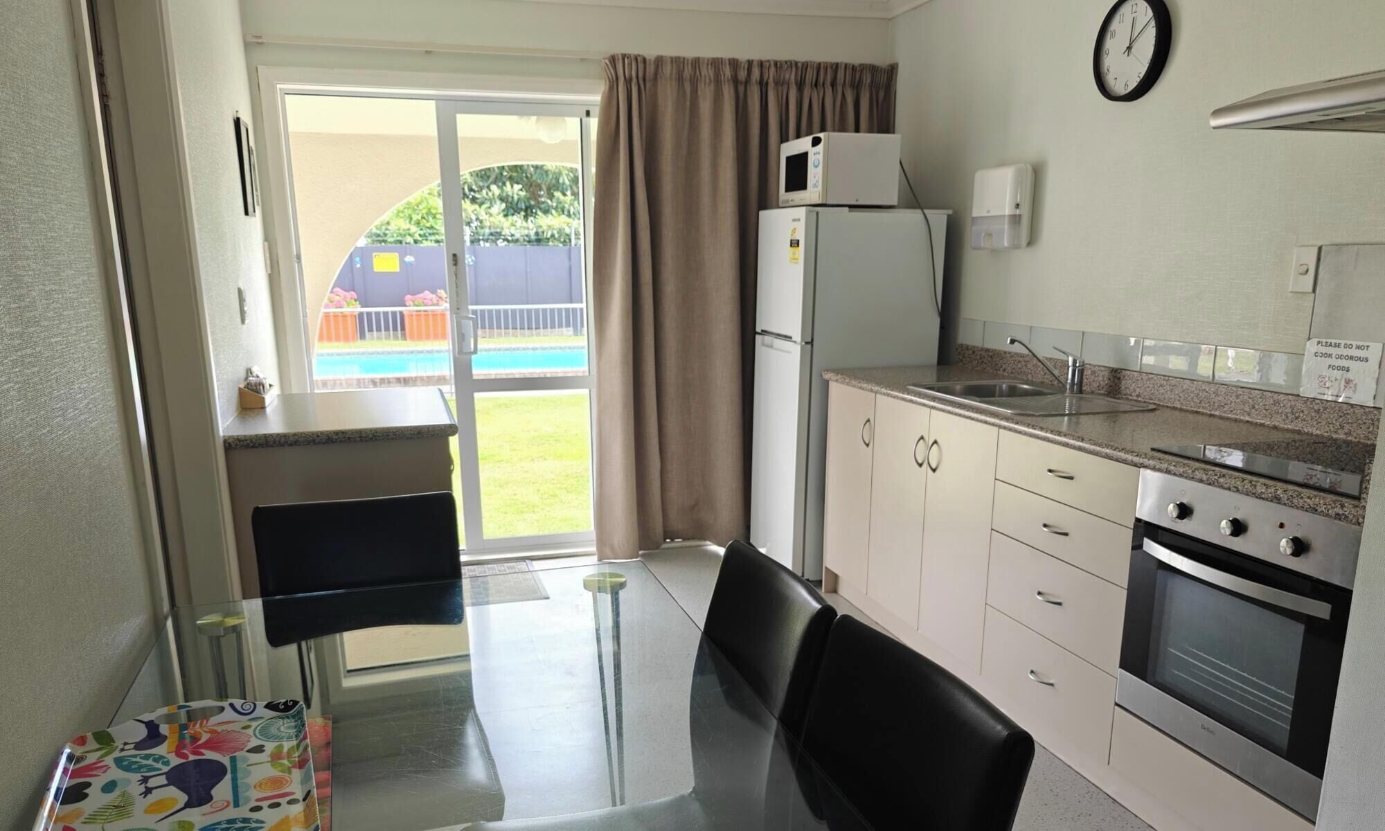 A compact kitchen with a refrigerator, microwave, stove, and sink. There's a dining table with chairs and a view of a pool outside.