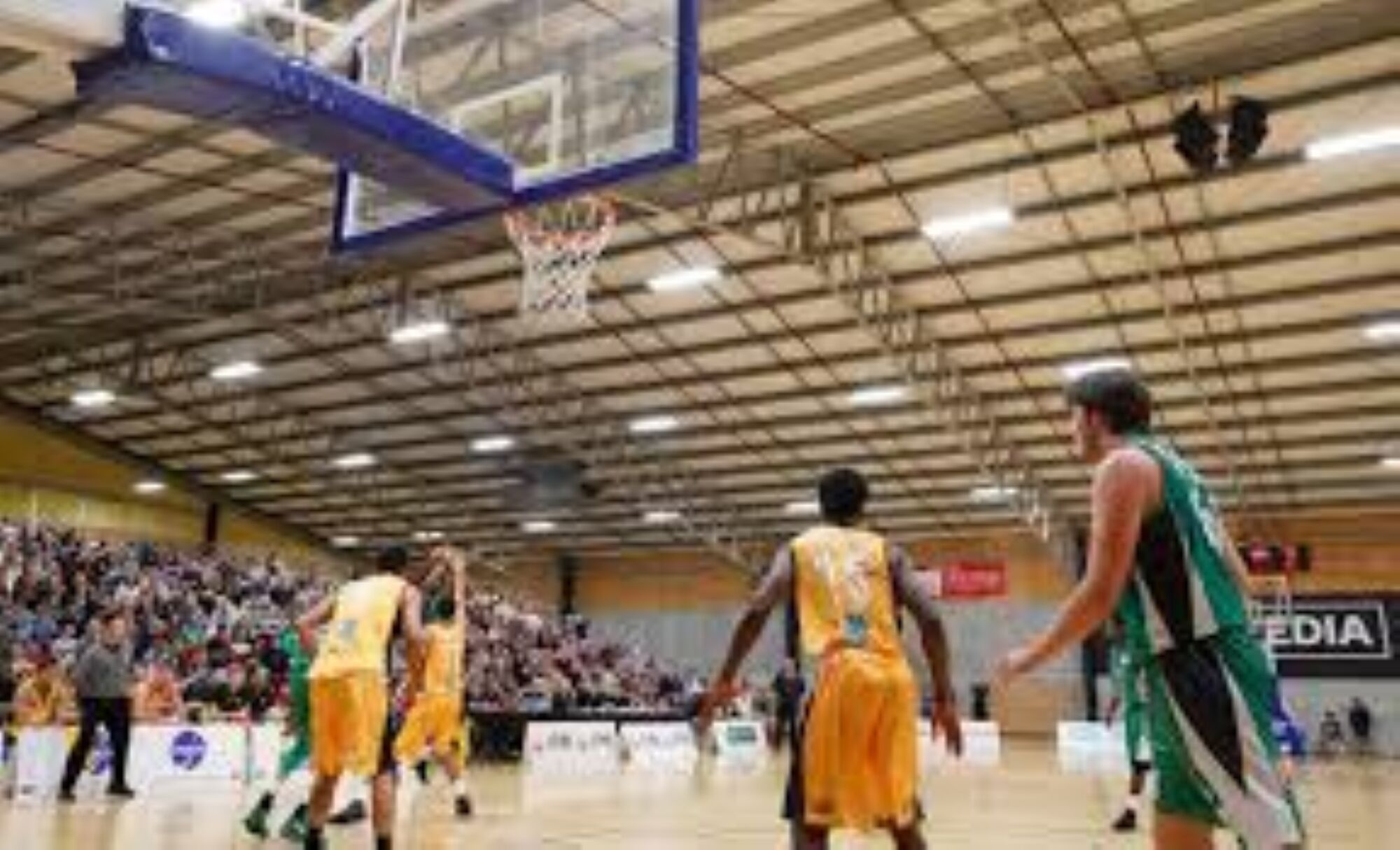 A basketball court with players in yellow and green uniforms, a visible basketball hoop, and spectators in the background.