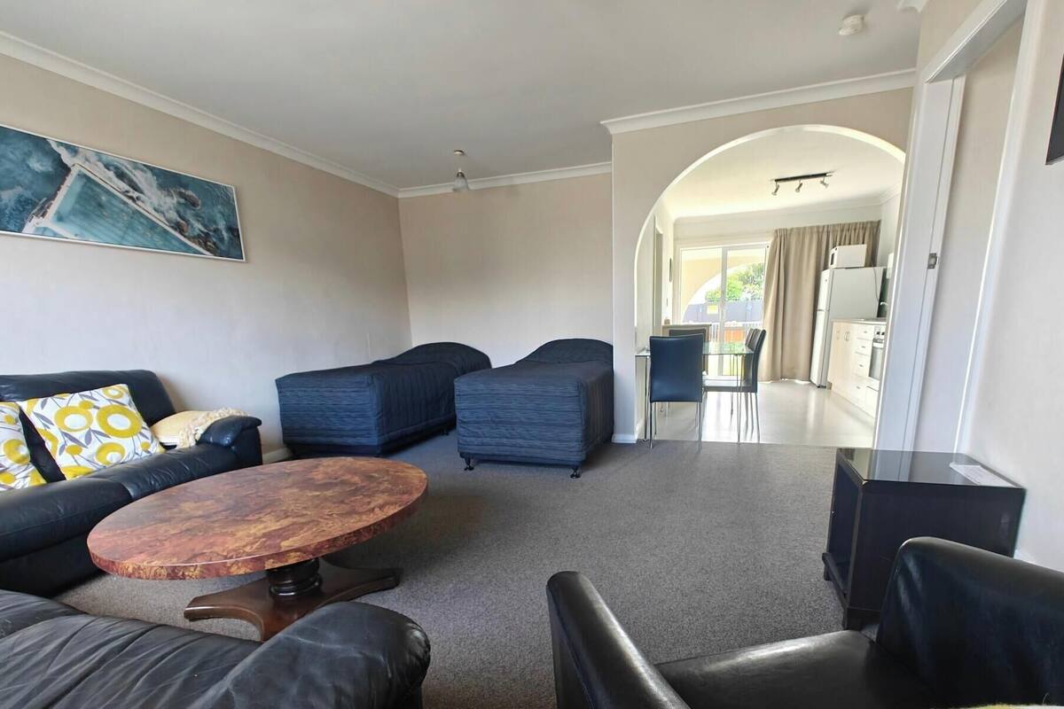 A modern living room with a sofa, two armchairs, a coffee table, and a painting on the wall. There is a kitchen visible through an archway.