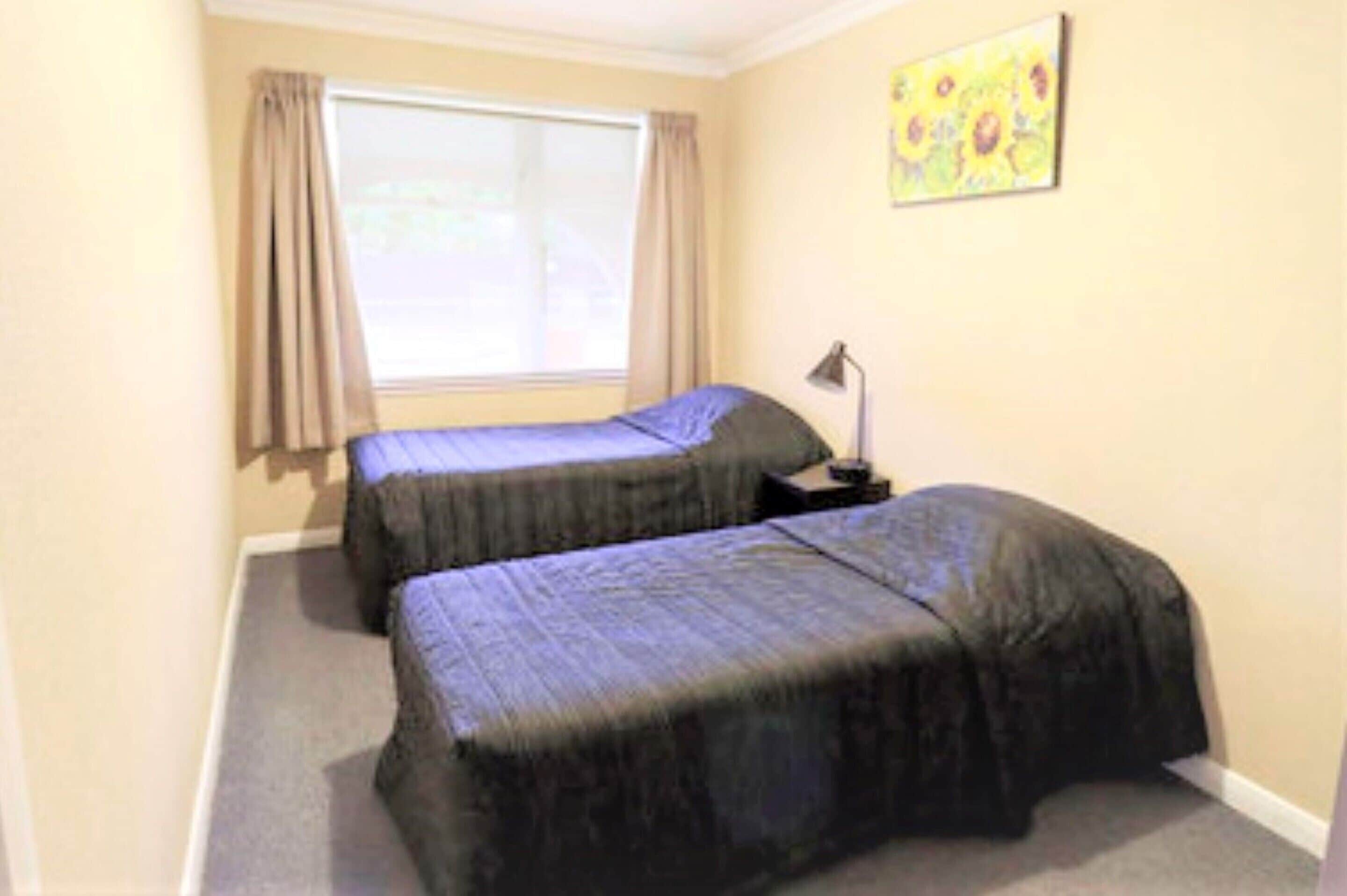 A small room with two beds, a window with curtains, a nightstand with a lamp, and a painting on the wall.