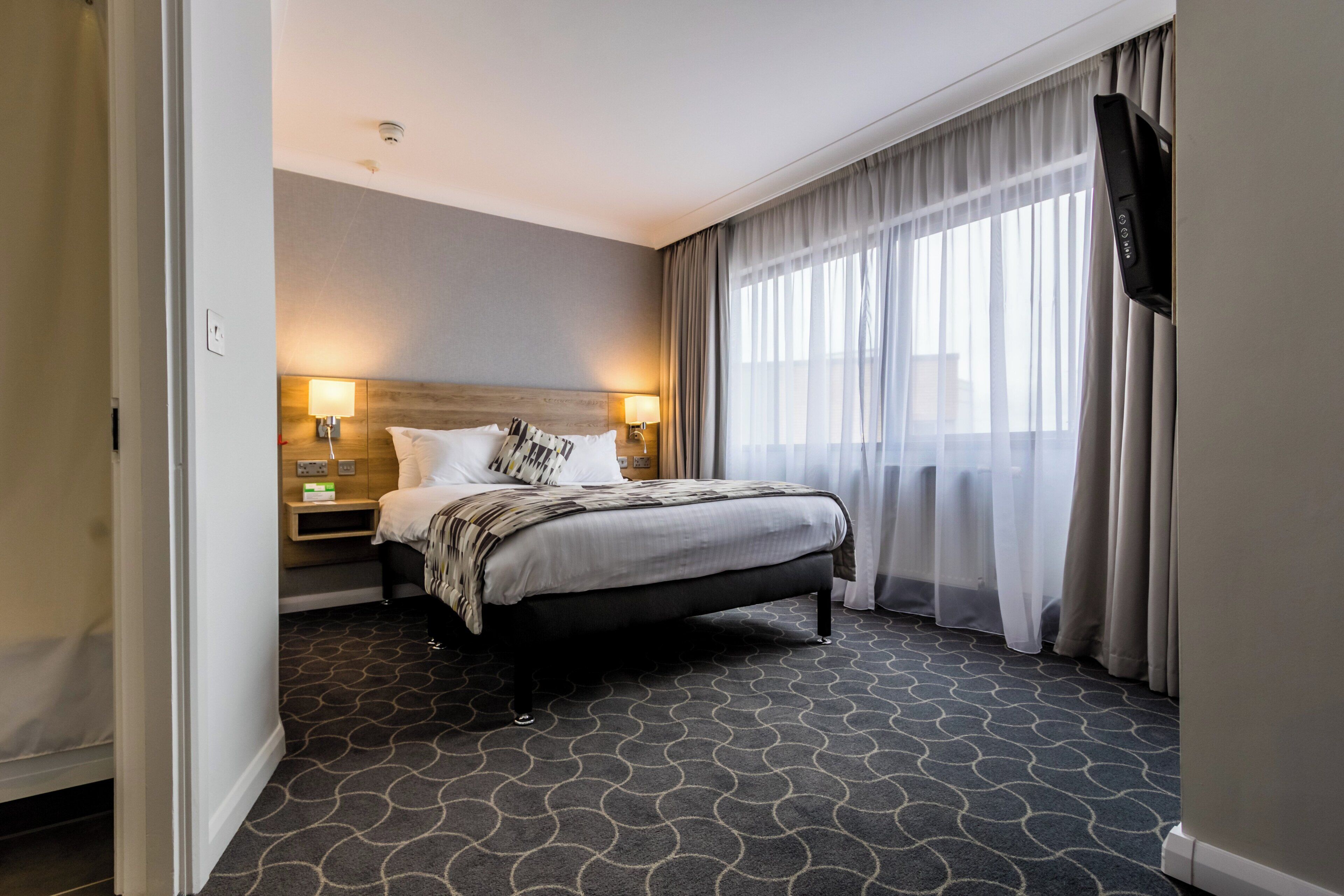 A hotel room with a bed, two bedside lamps, a TV, and a window with curtains.