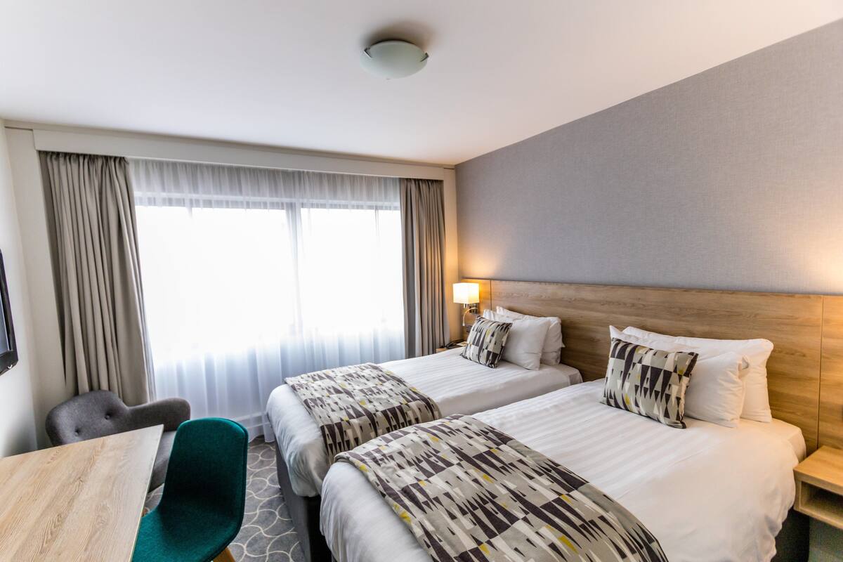 A hotel room with two beds, a desk, a chair, and a window with curtains.
