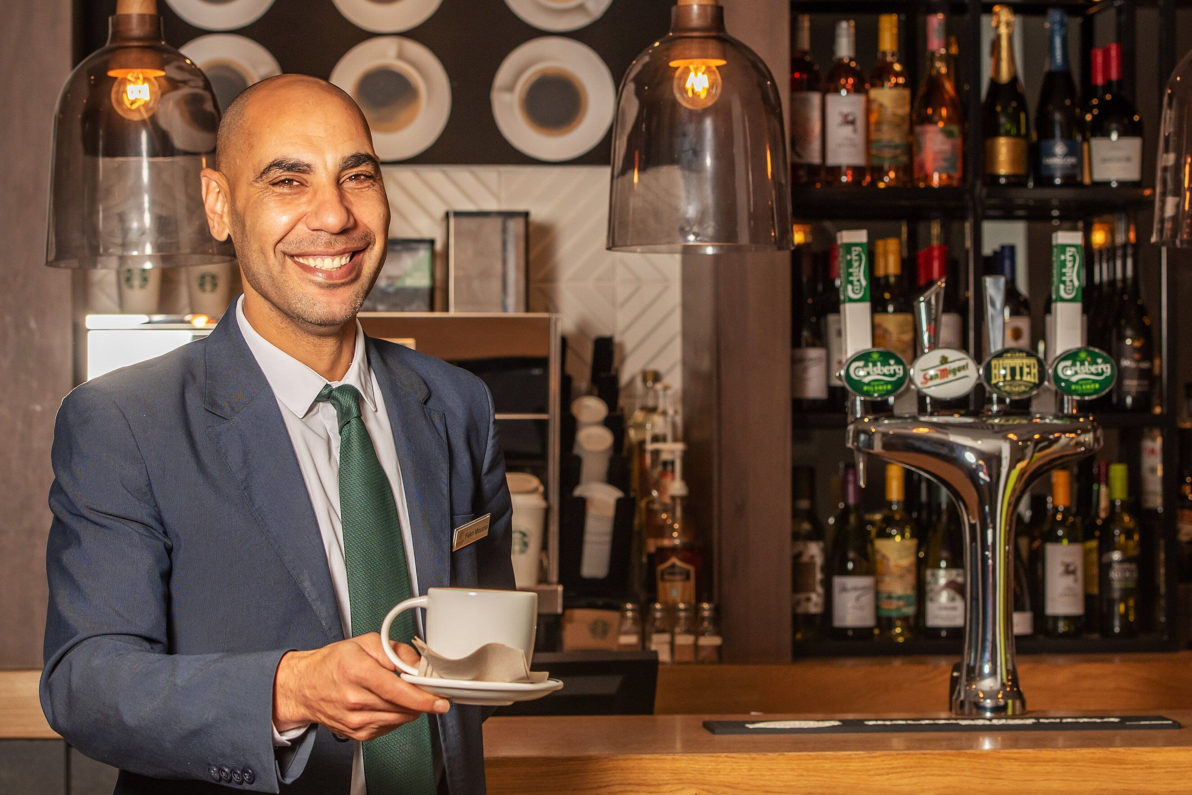 A bar area with a person holding a cup, a bar tap, and shelves with bottles.