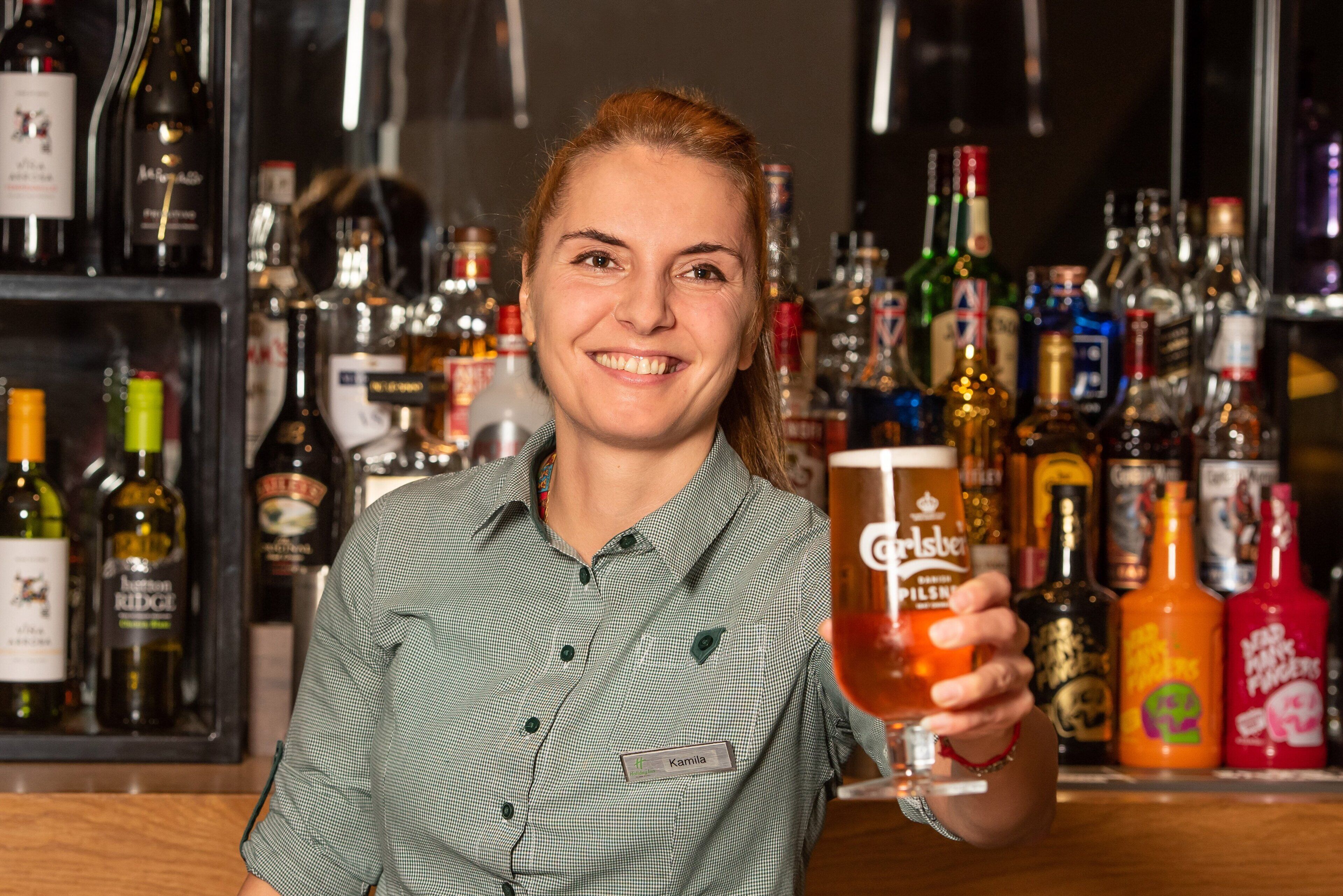A person holding a glass of beer at a bar with various bottles of alcohol in the background.
