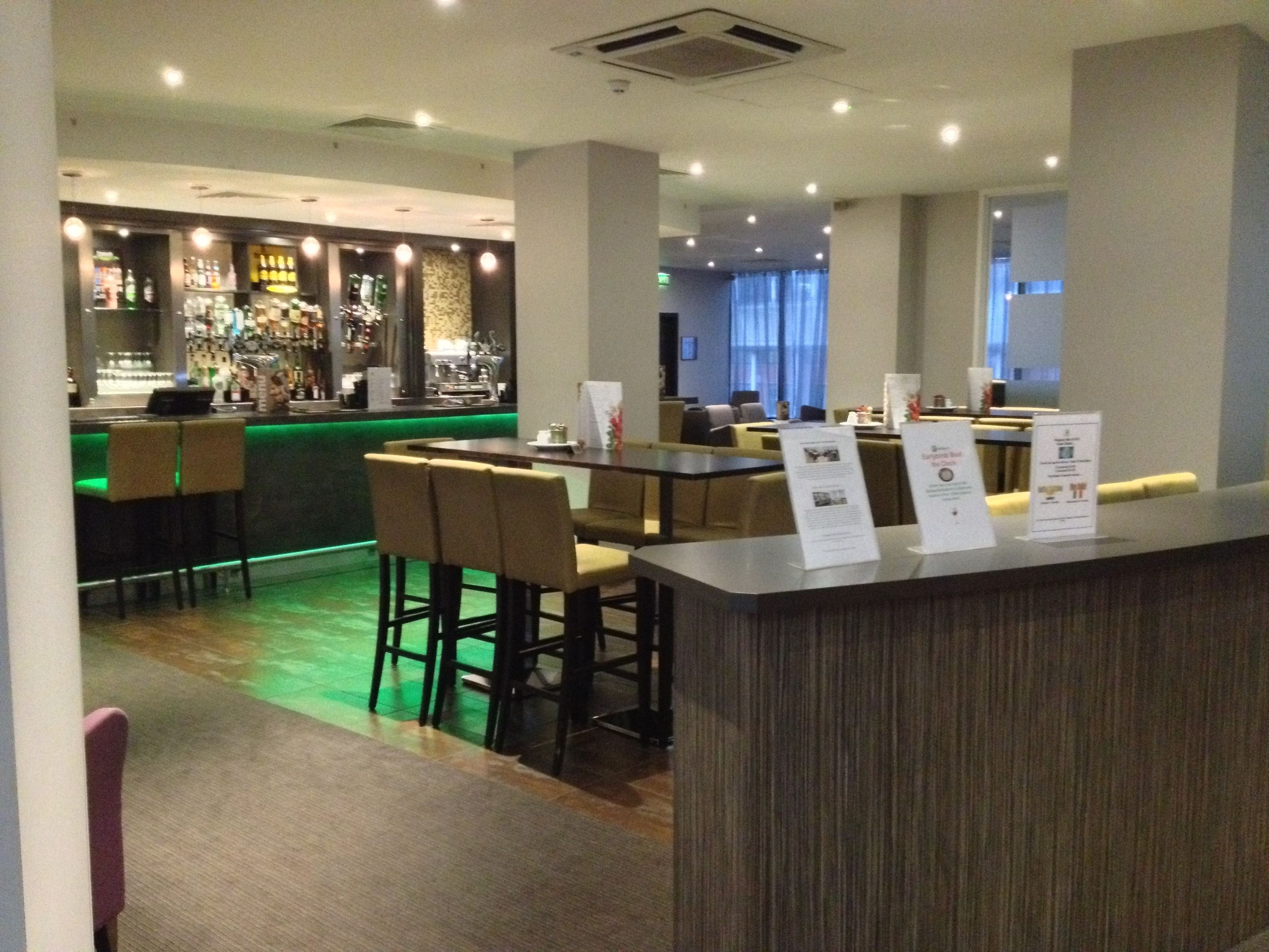 Eine moderne Bar mit grüner Beleuchtung, Sitzgelegenheiten mit hoher Rückenlehne und Informationsschildern auf der Theke.