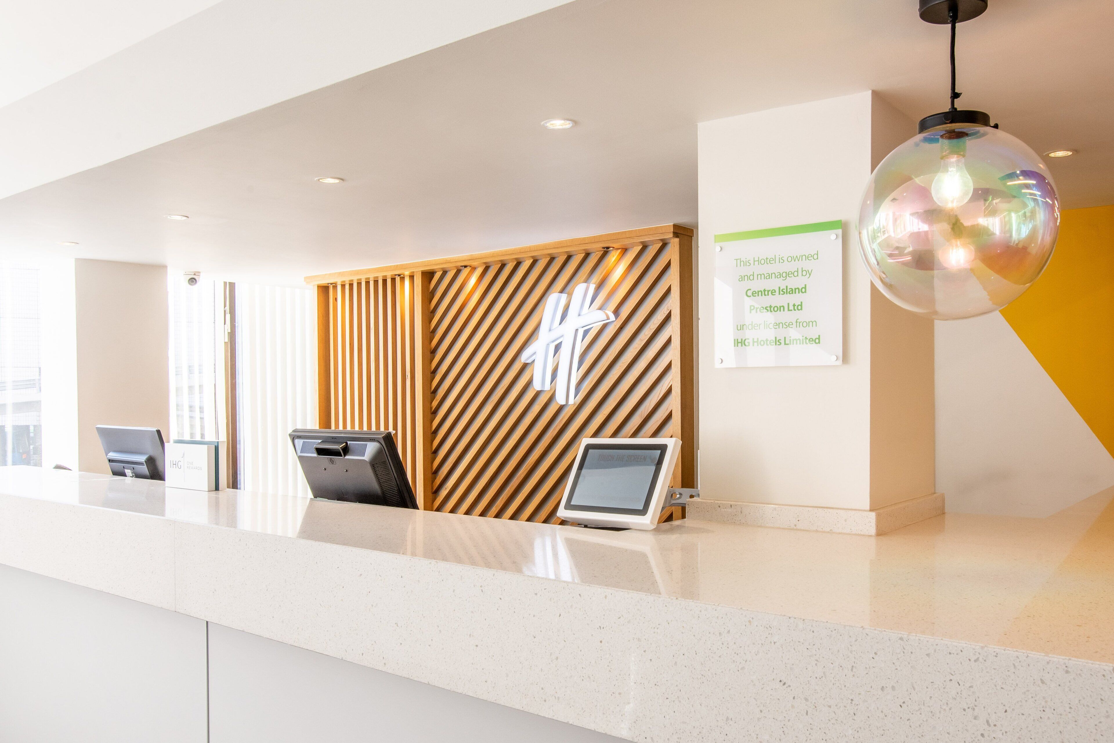 A hotel reception area with a modern design, featuring a reception desk, a wooden panel wall with a logo, a digital information board, and a unique spherical pendant light.