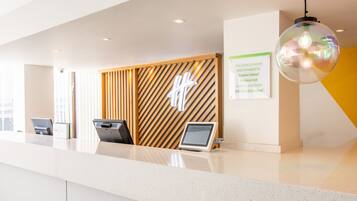 A hotel reception area with a modern design, featuring a reception desk, a wooden panel wall with a logo, a digital information board, and a unique spherical pendant light.