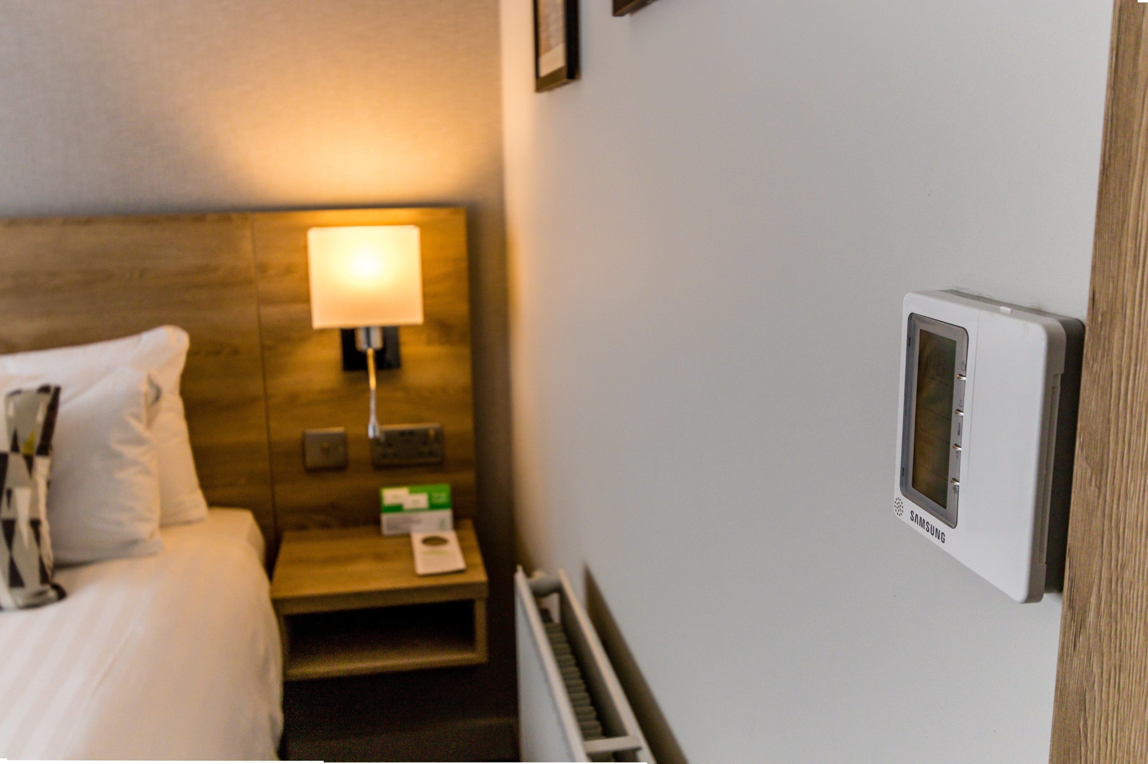 Una habitación de hotel con cama, una mesita de noche con lámpara, un termostato y un cuadro en la pared.