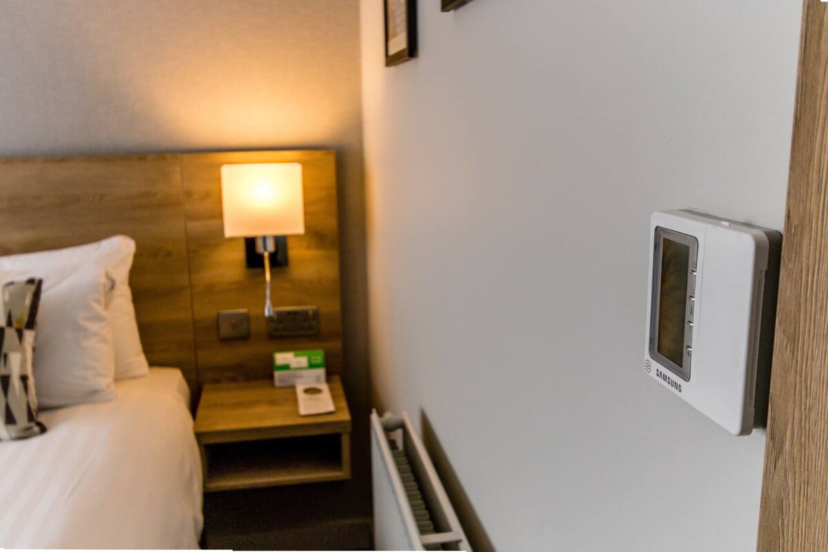 A hotel room with a bed, a bedside table with a lamp, a thermostat, and a framed picture on the wall.