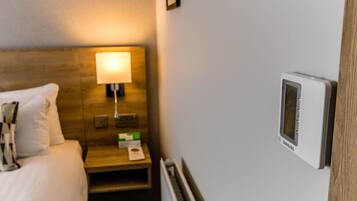 Una habitación de hotel con cama, una mesita de noche con lámpara, un termostato y un cuadro en la pared.