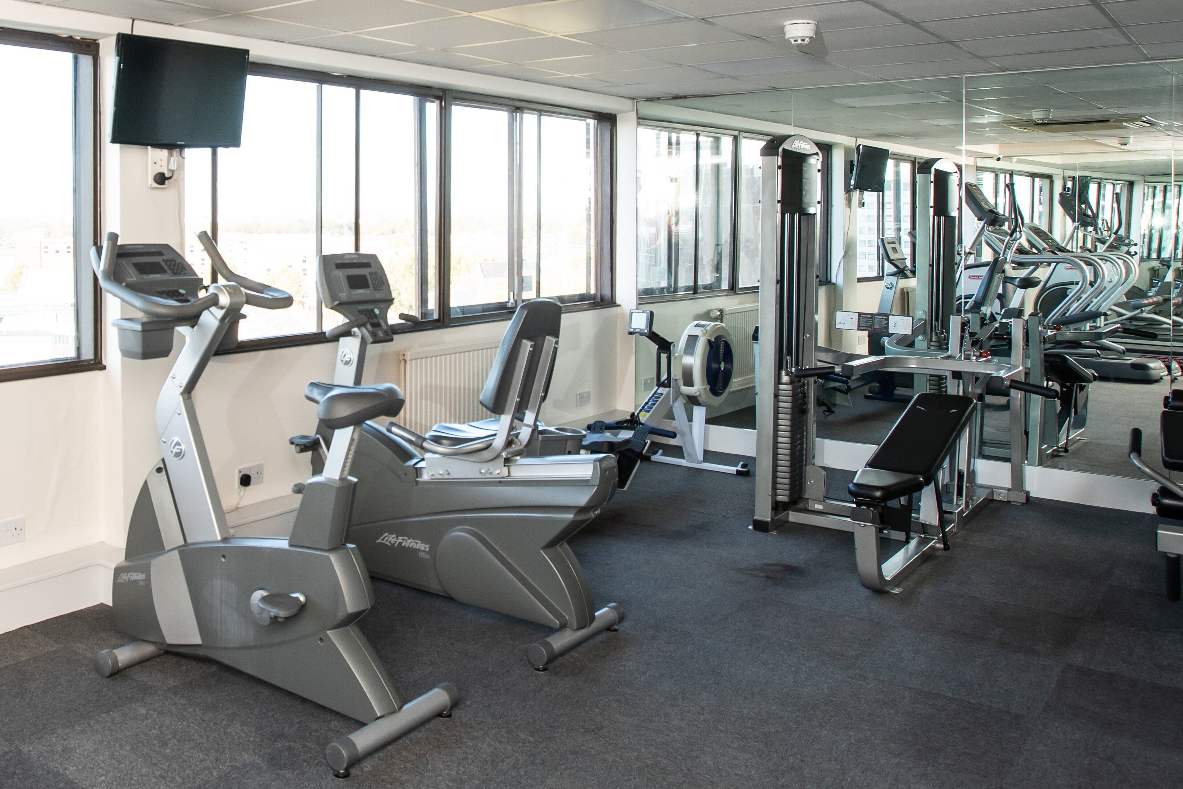 Uma academia com vários equipamentos de exercício, janelas amplas e um espelho grande.