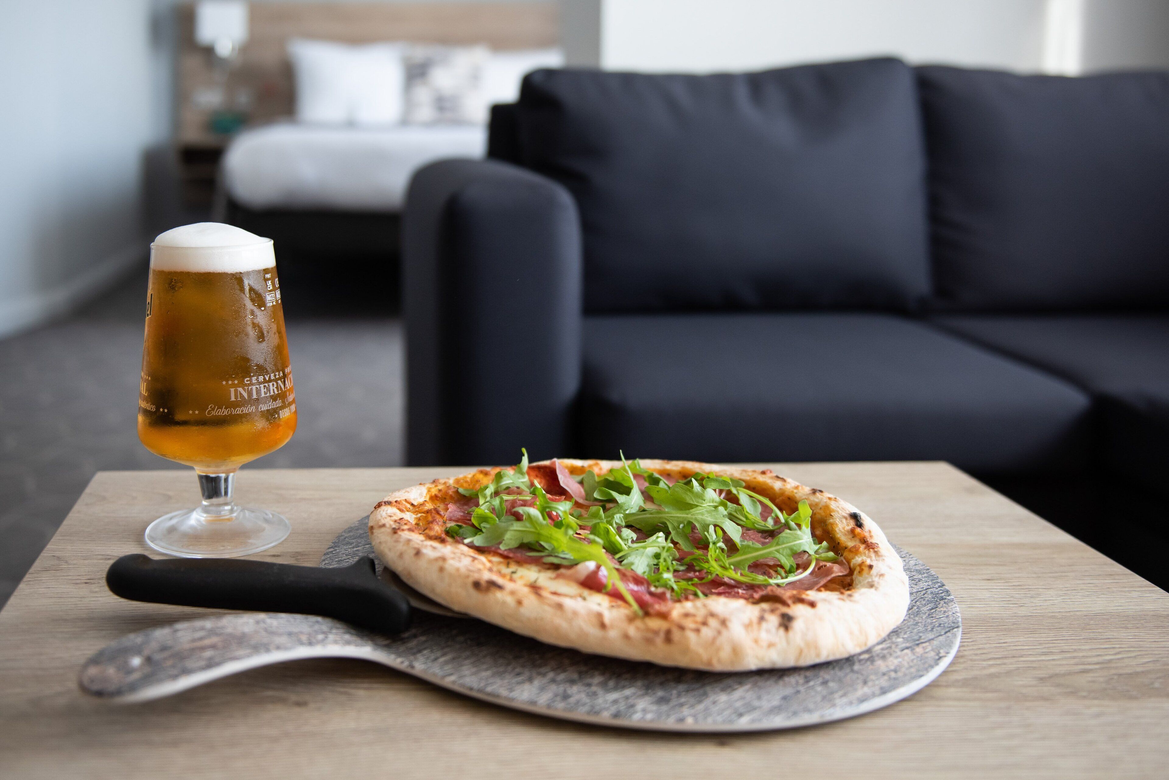 A pizza on a wooden table with a pizza cutter beside it, and a glass of beer in the background.