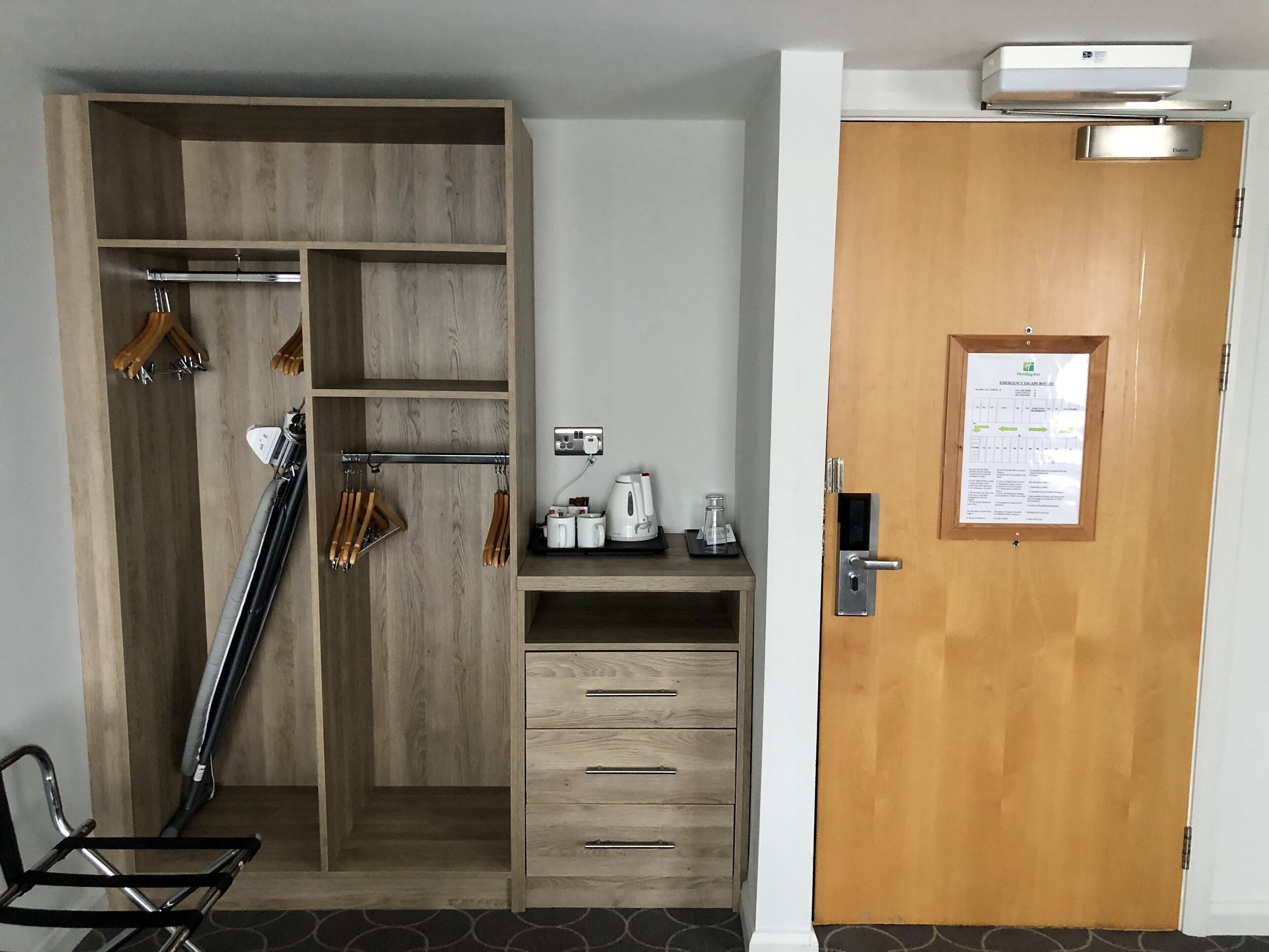 A hotel room with a wooden wardrobe, a desk with a kettle, and a door with a posted notice.
