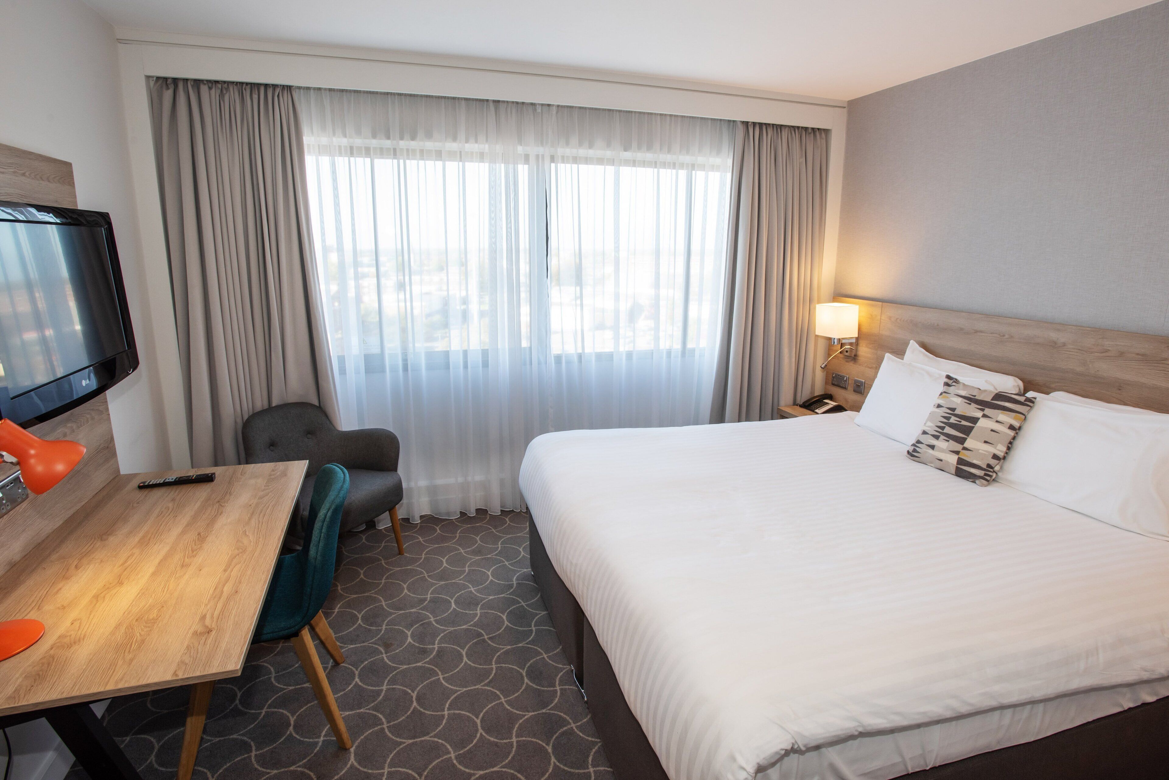 A hotel room with a bed, a desk, a chair, a TV, and a window with curtains.