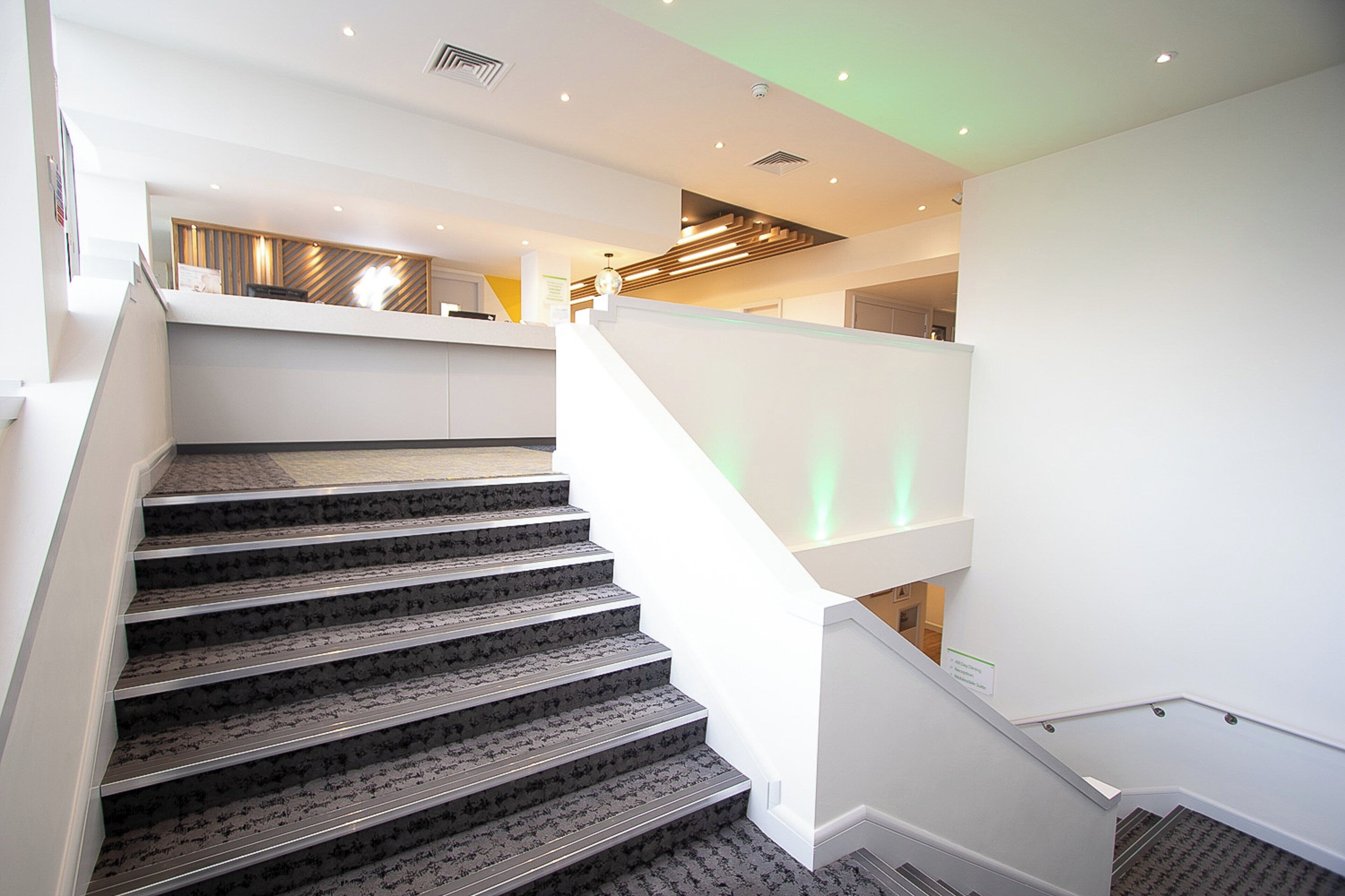 Una escalera moderna con alfombra estampada y barandillas metálicas, que conduce a un rellano en el segundo piso con baranda de vidrio.