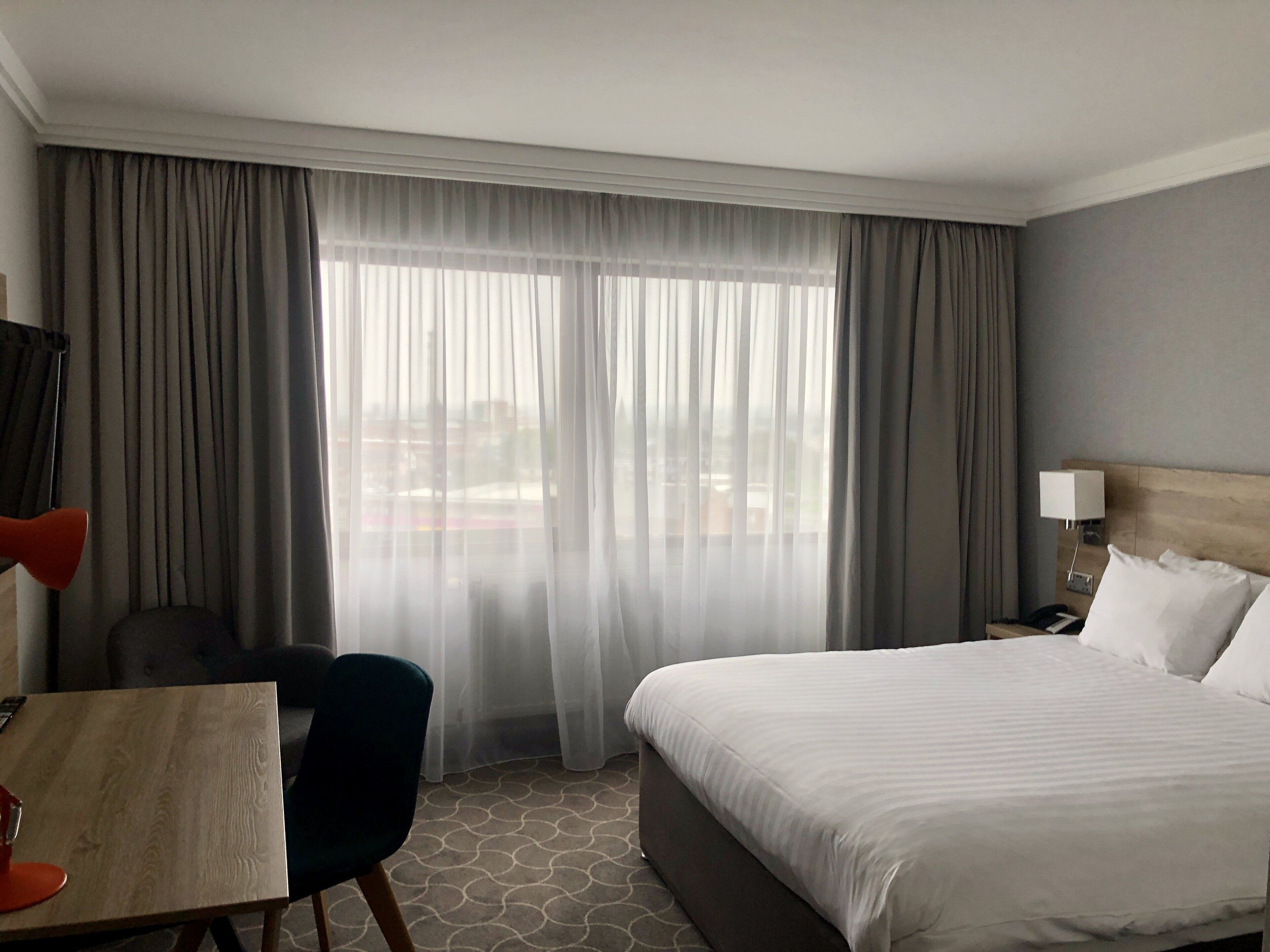 Une chambre d’hôtel avec un lit, un bureau, une chaise, une lampe et une vue par la fenêtre.