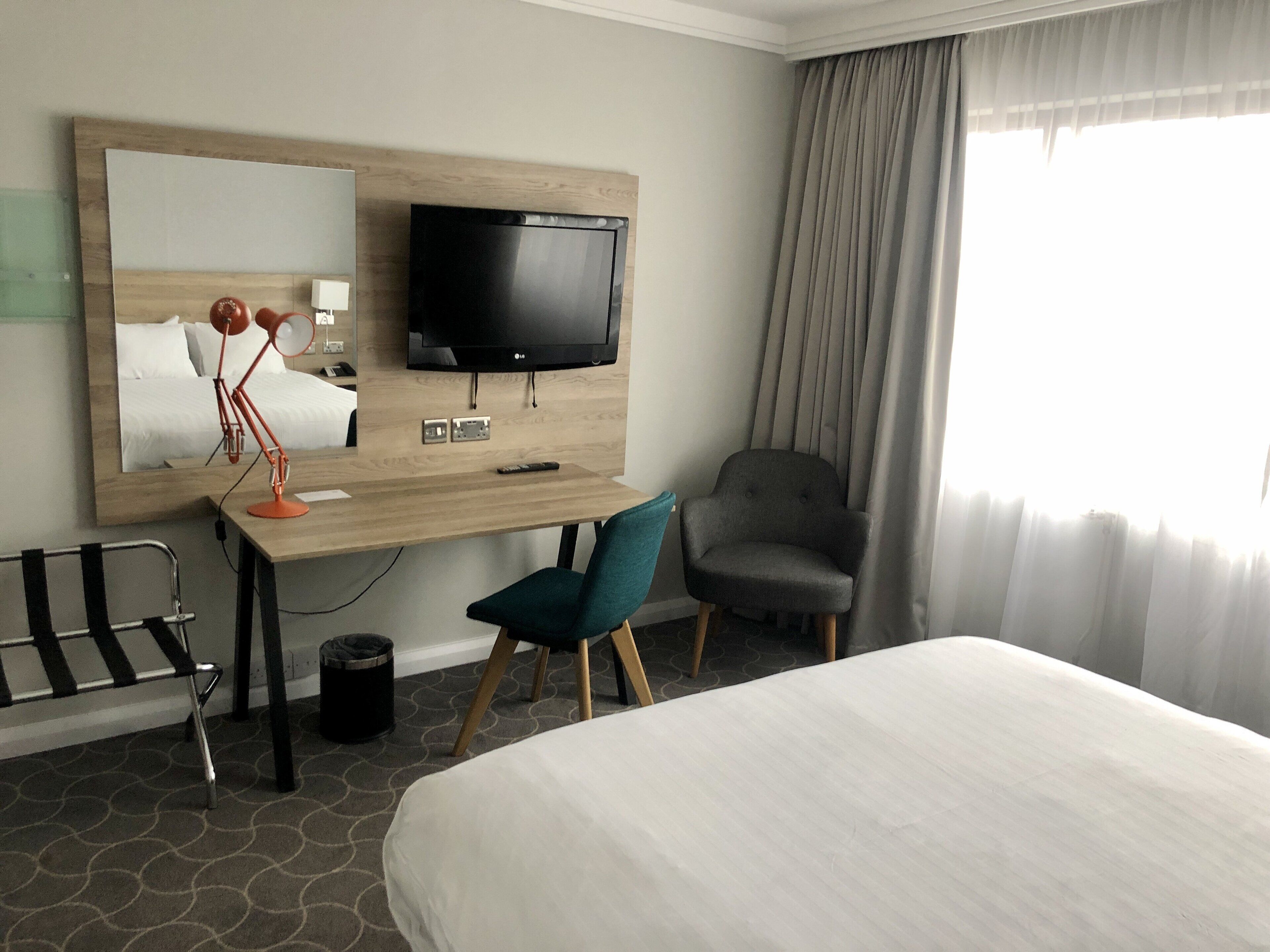 A hotel room with a bed, a desk with a TV, a chair, and a large mirror.