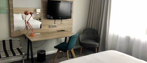 A hotel room with a bed, a desk with a TV, a chair, and a large mirror.