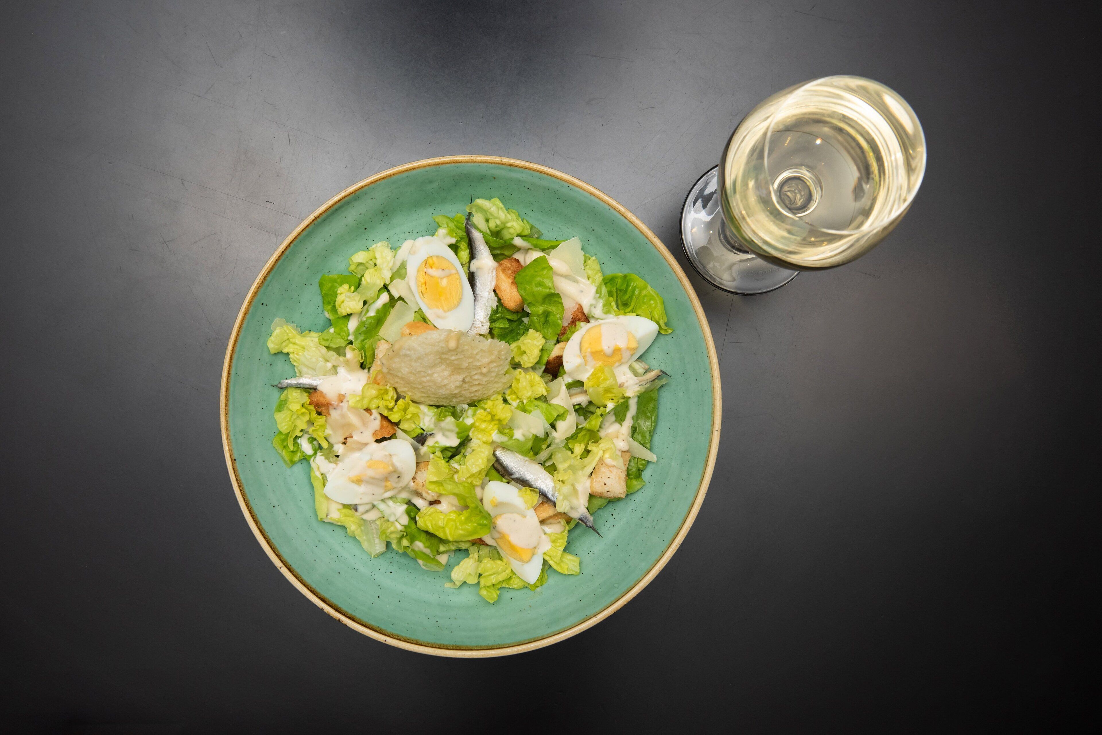 Une assiette verte contenant une salade composée de laitue, d’œufs et d’un morceau de pain, accompagnée d’un verre de vin blanc, le tout posé sur une surface sombre.
