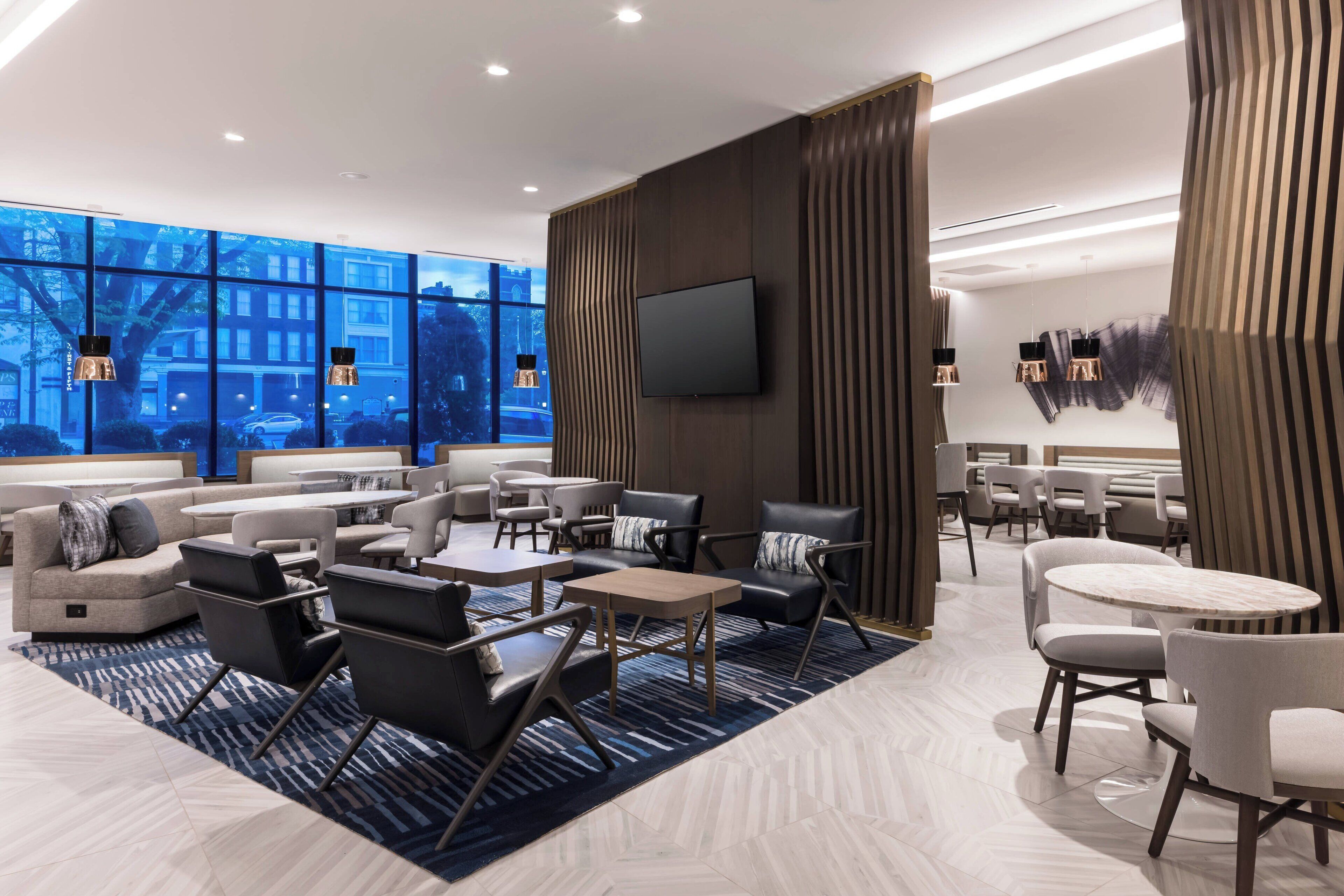 A modern hotel lobby with a large seating area, a flat-screen TV mounted on the wall, and a view of the city through large windows.