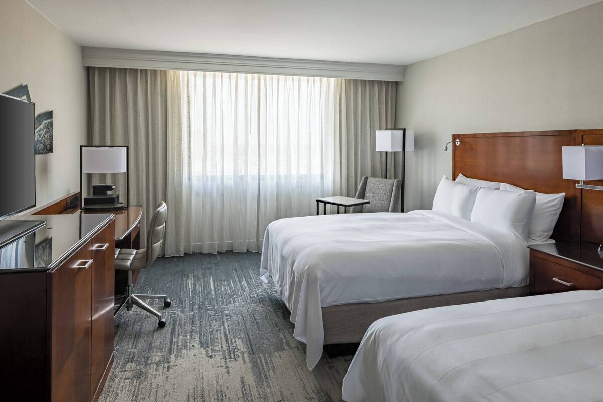 A hotel room with two beds, a desk, a chair, a TV, and a large window with curtains.