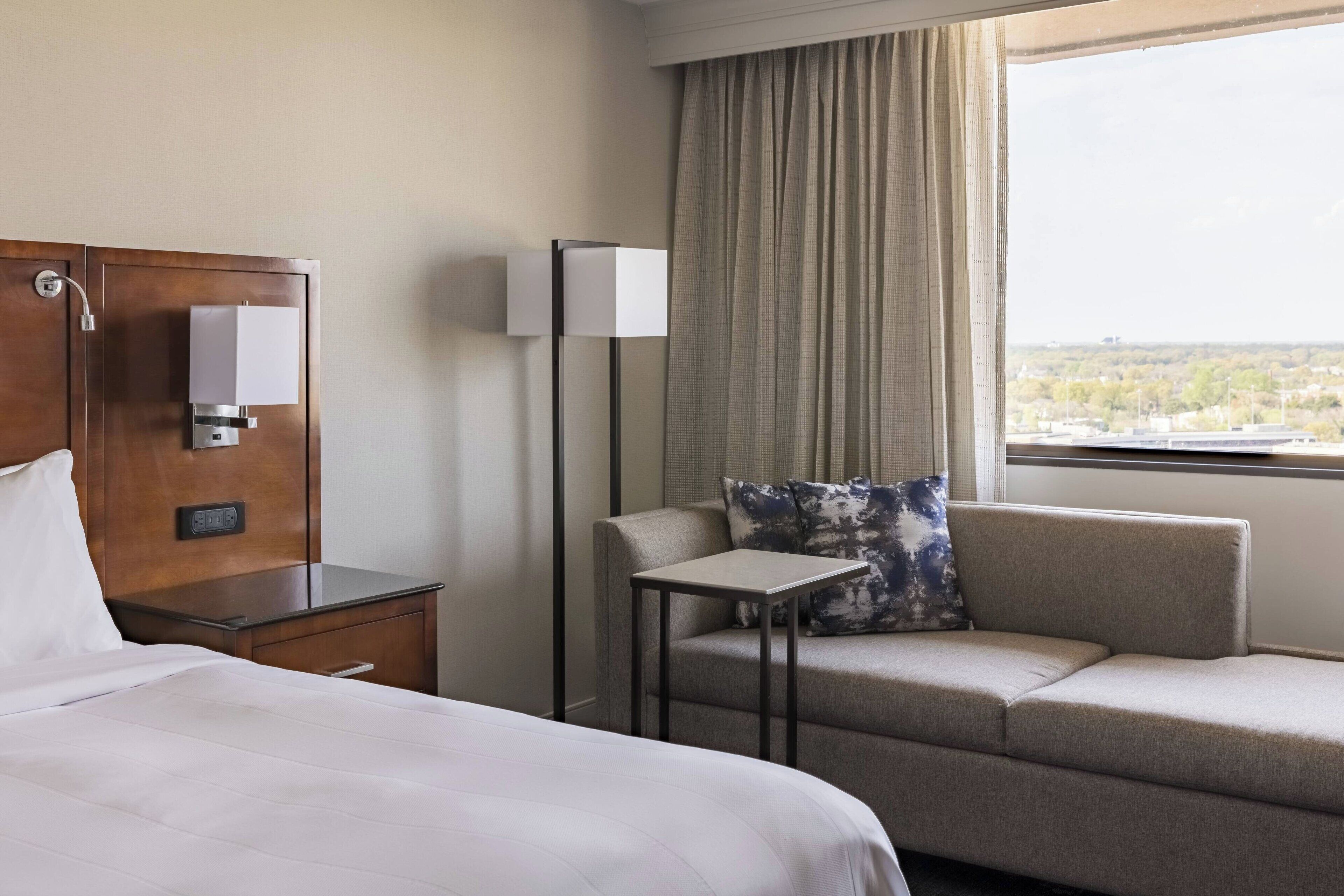 Ein Hotelzimmer mit Bett, Schreibtisch, Sofa, einem kleinen Tisch, einer Lampe und einem Fenster mit Aussicht.