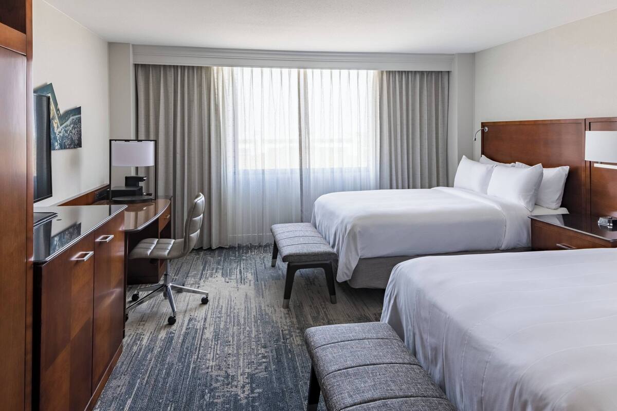 A hotel room with two beds, a desk, a chair, and a large window with curtains.