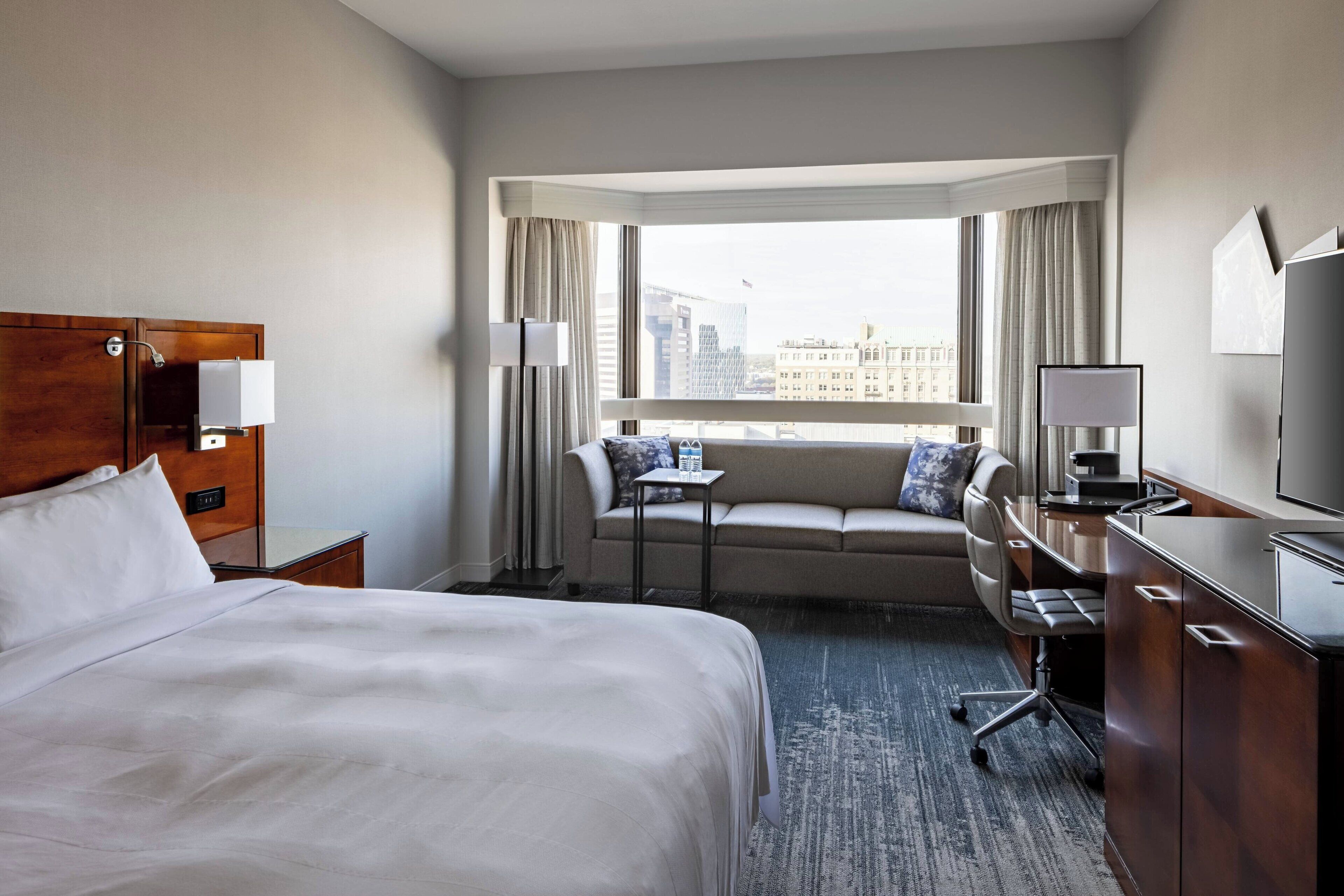 A hotel room with a large bed, a desk with a computer, a sofa, a small table, and a window with a city view.