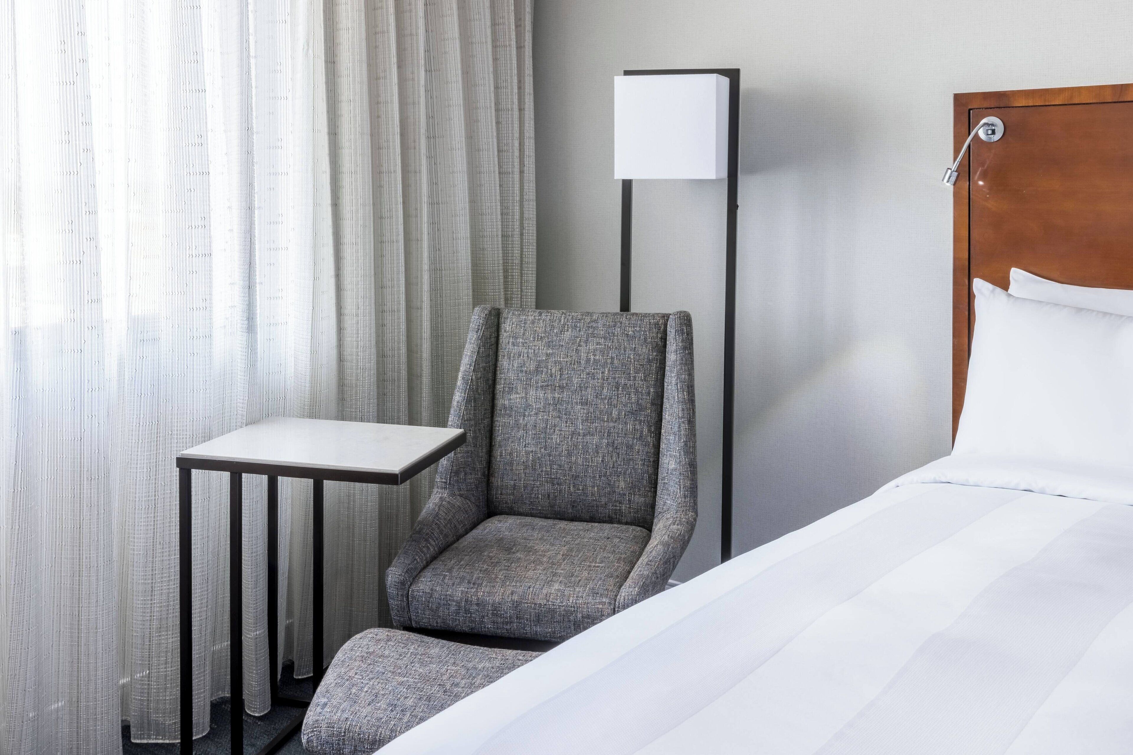 Habitación de hotel con cama, un sillón gris, una mesita, una lámpara de pie y una ventana con cortinas.