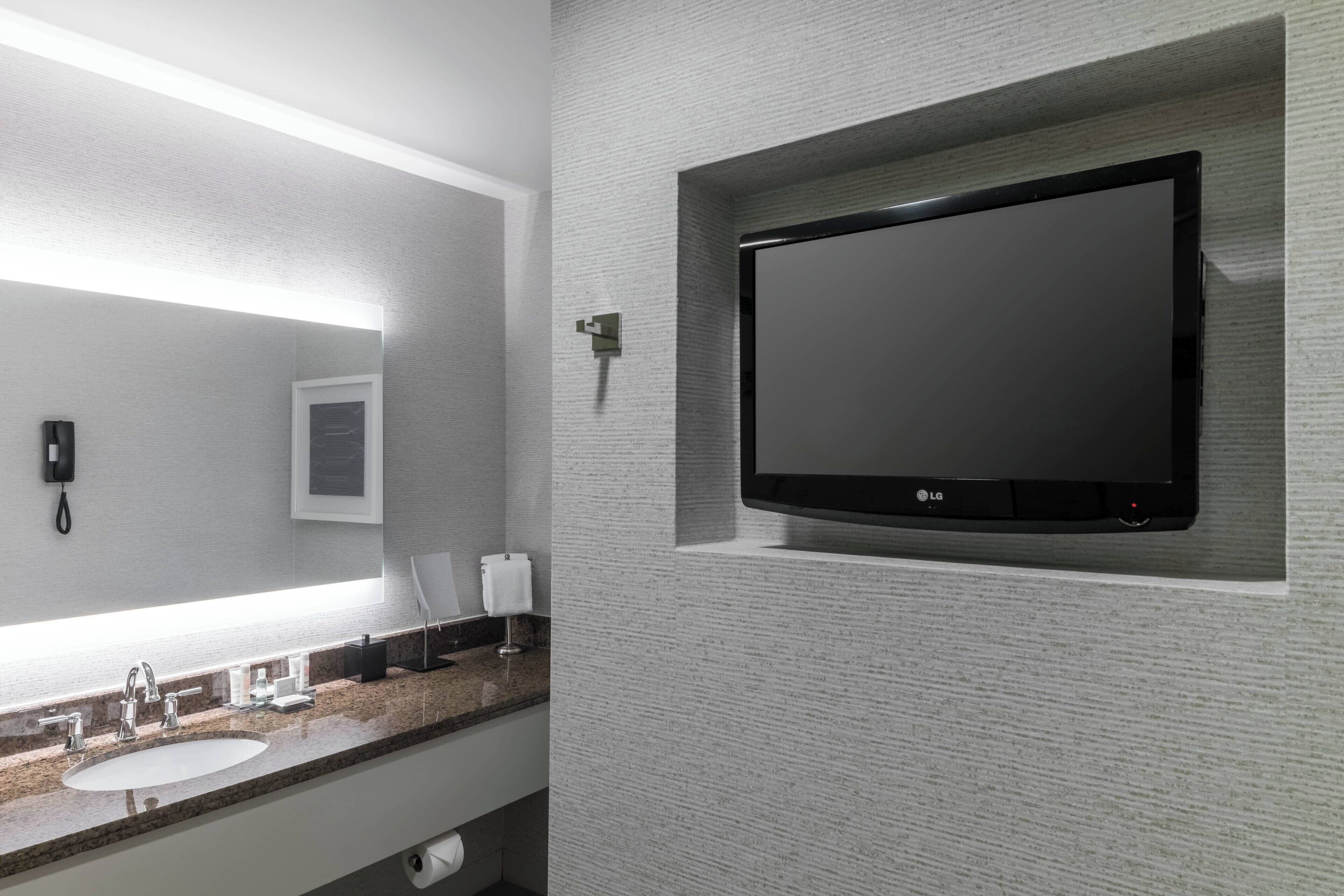 Een moderne badkamer met een granieten aanrechtblad, een witte wastafel, een spiegel, een zwarte LG TV aan de muur en een telefoon.