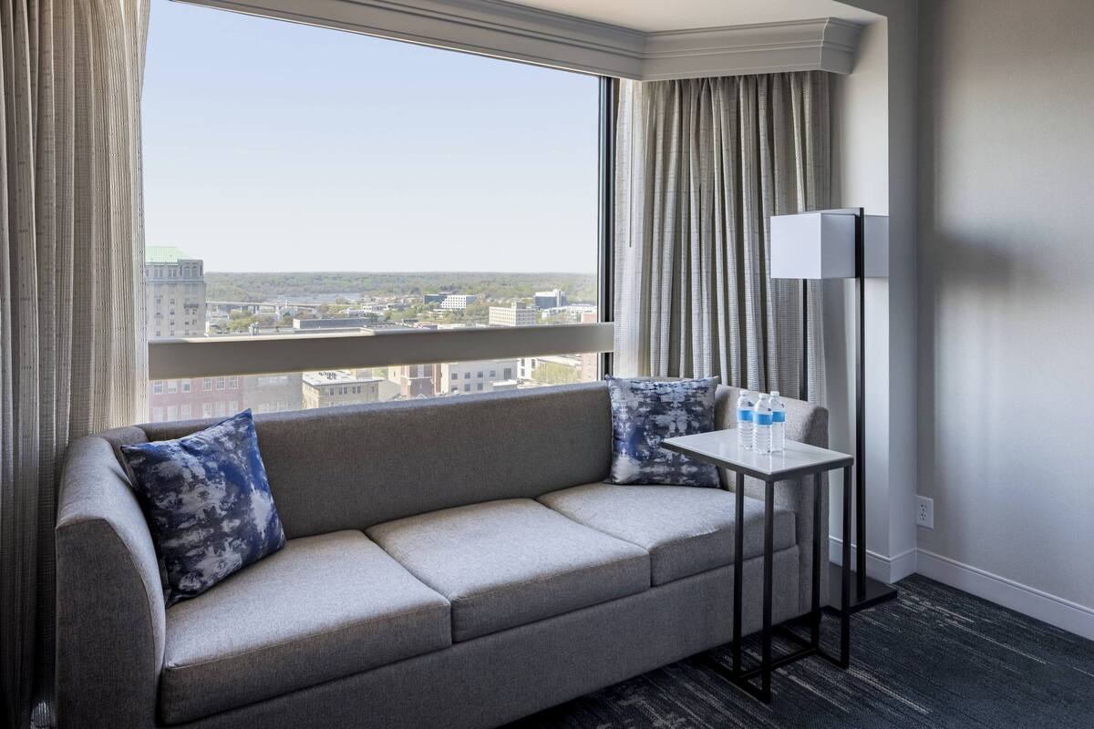 A modern hotel room with a grey sofa, two patterned pillows, a small table with water bottles, and a view of the city through large windows.