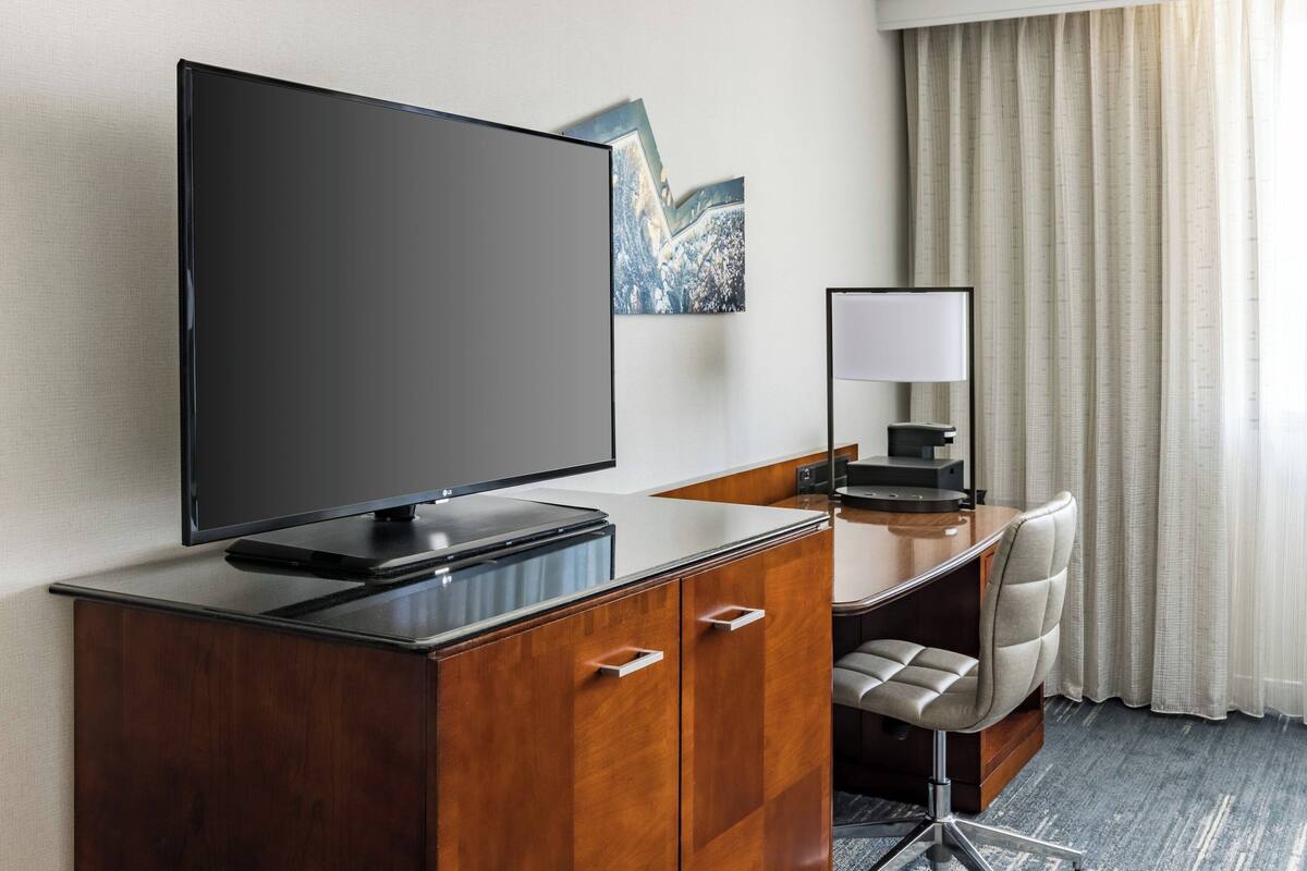 A hotel room with a flat-screen TV, a desk with a chair, a mirror, and a window with curtains.