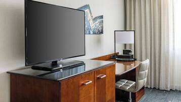 A hotel room with a flat-screen TV, a desk with a chair, a mirror, and a window with curtains.