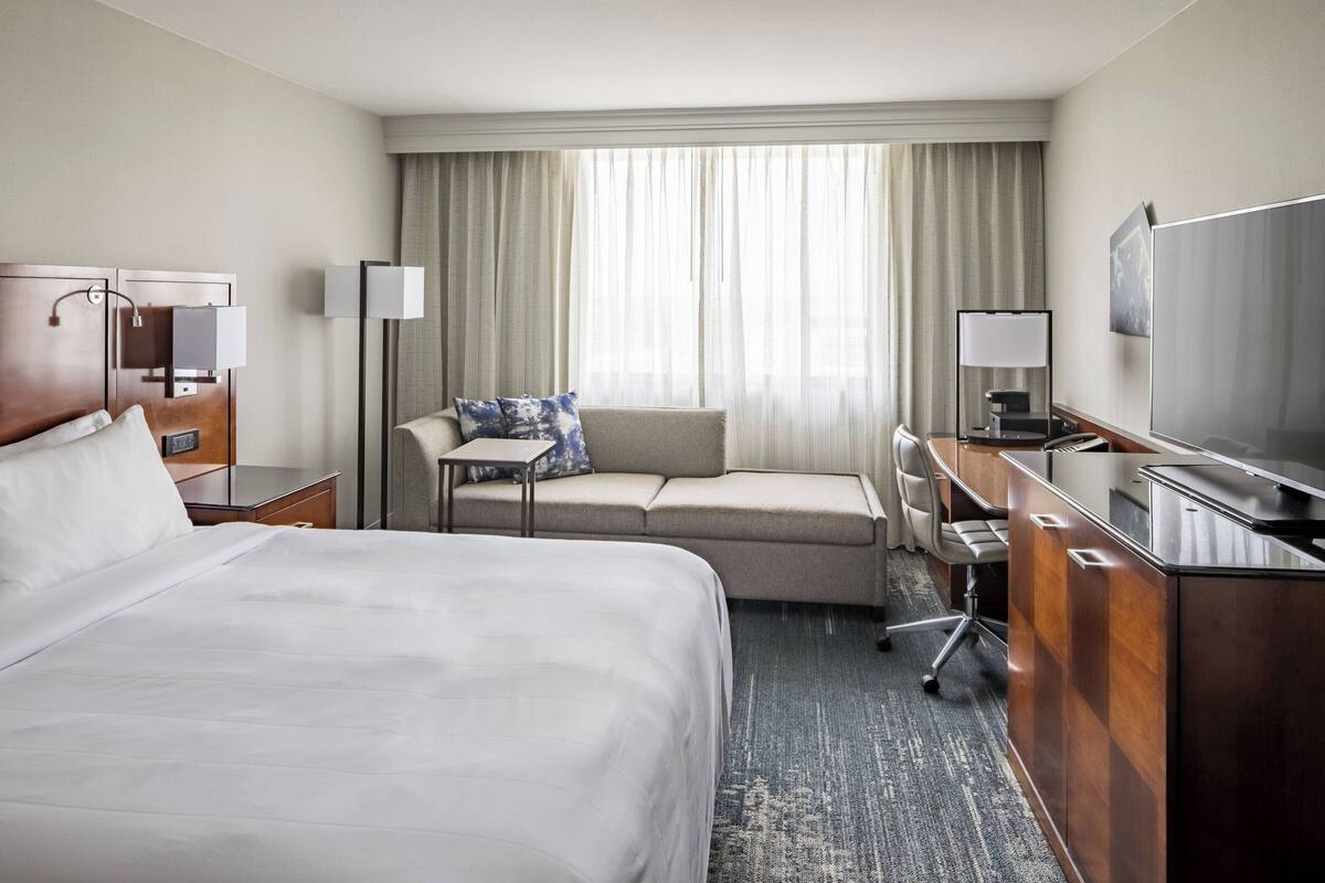 A hotel room with a large bed, a sofa, a desk with a computer, a TV, and a window with curtains.