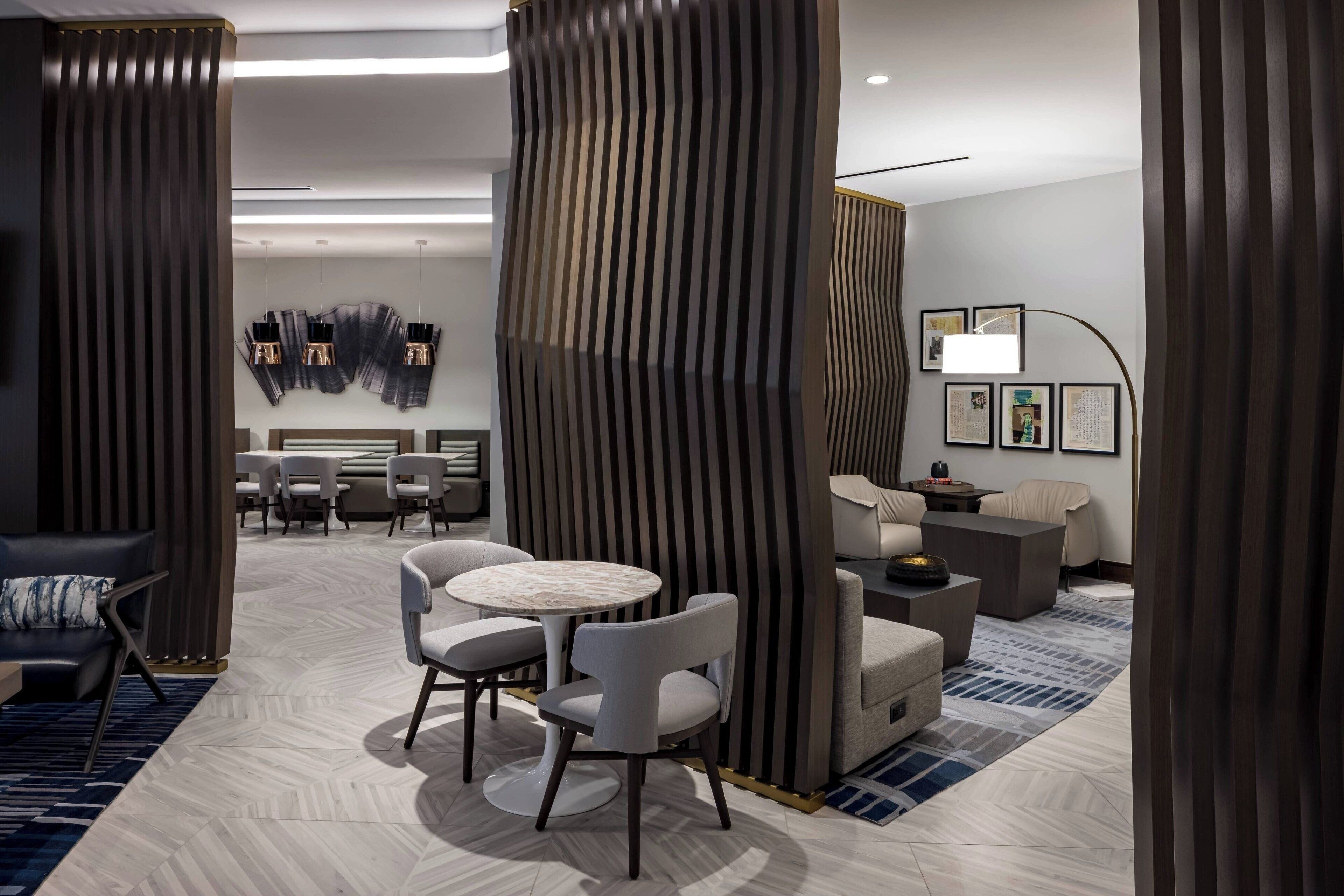 A modern hotel room with a sofa, two armchairs, a round table, and a dining area with a chandelier.
