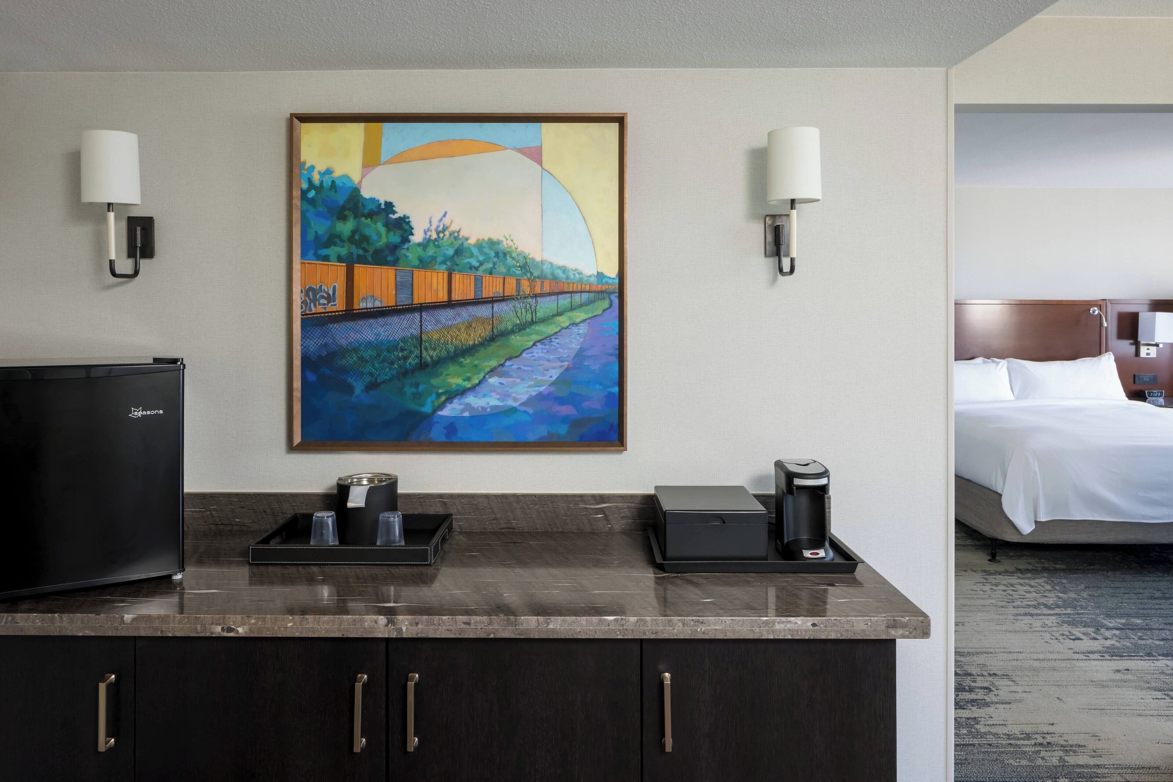 A hotel room with a bed, a desk with a coffee maker, a TV, and a painting on the wall.