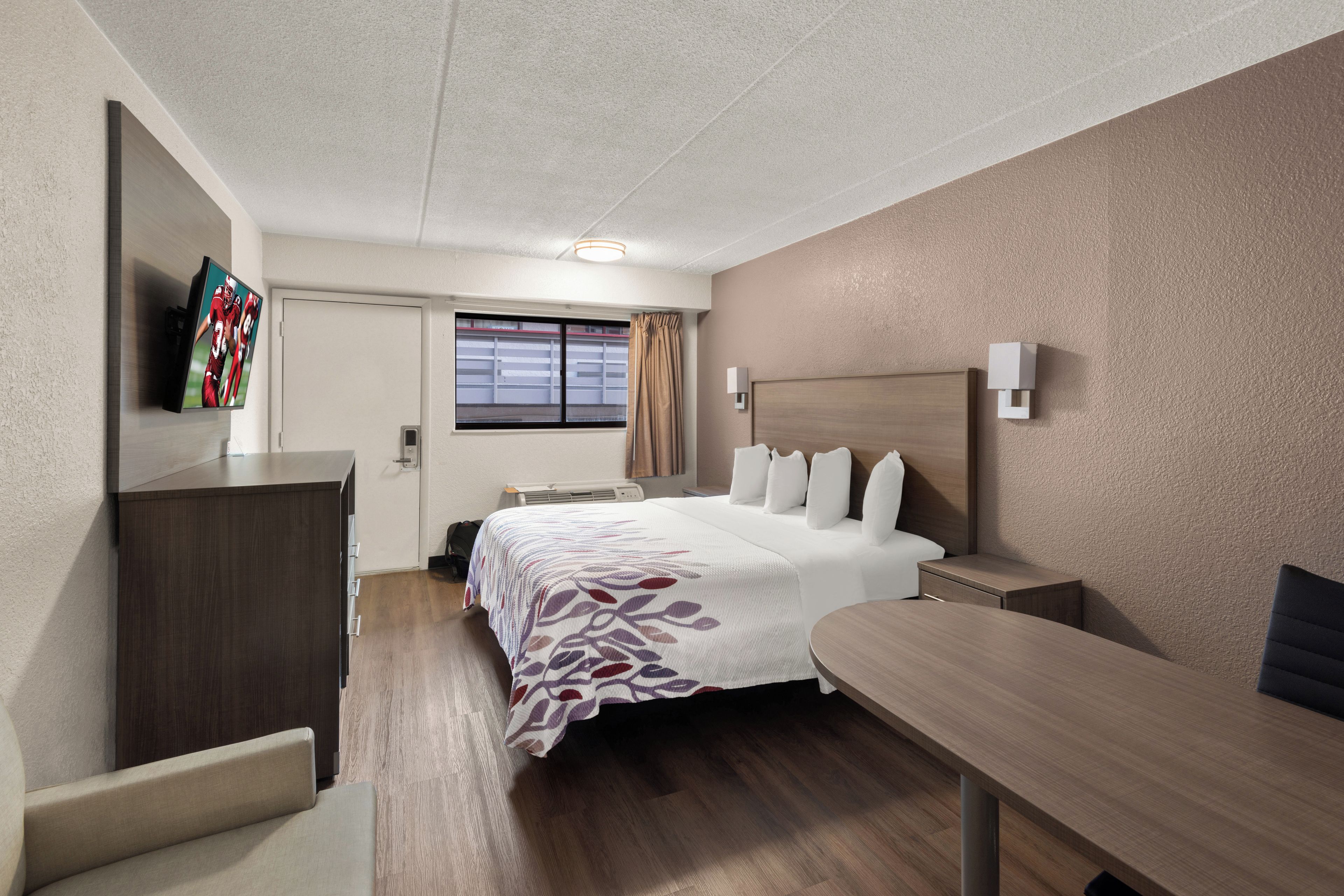 Ein Hotelzimmer mit Bett, Nachttisch, Schreibtisch, Sessel, Wandfernseher und einem Vorhangfenster.