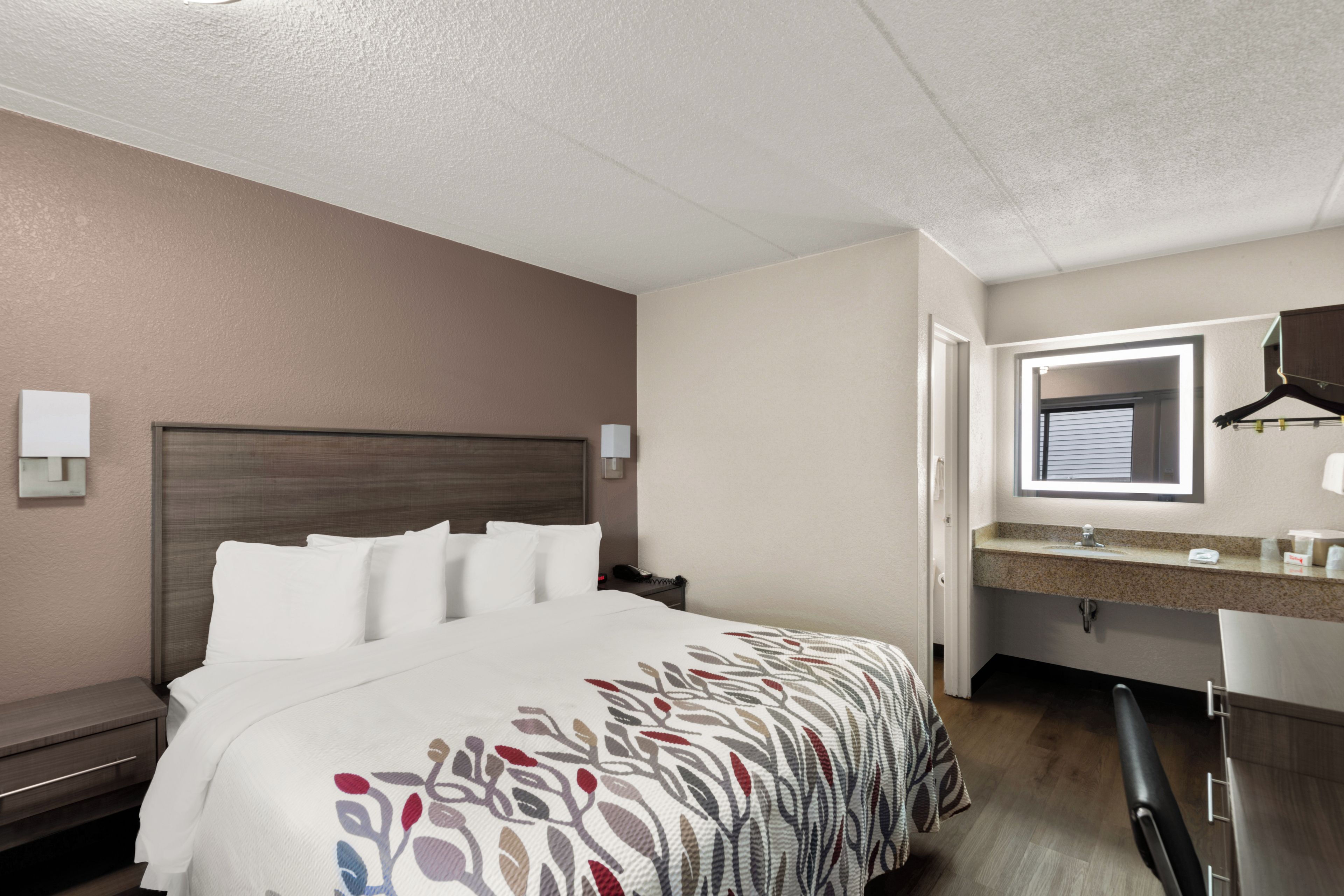 A hotel room with a bed, bedside table, nightstand, and a bathroom with a sink and mirror.