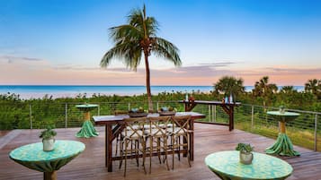 Terraza de madera con mesa y sillas, con vista a una playa y al océano.
