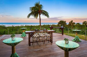 Terraza de madera con mesa y sillas, con vista a una playa y al océano.