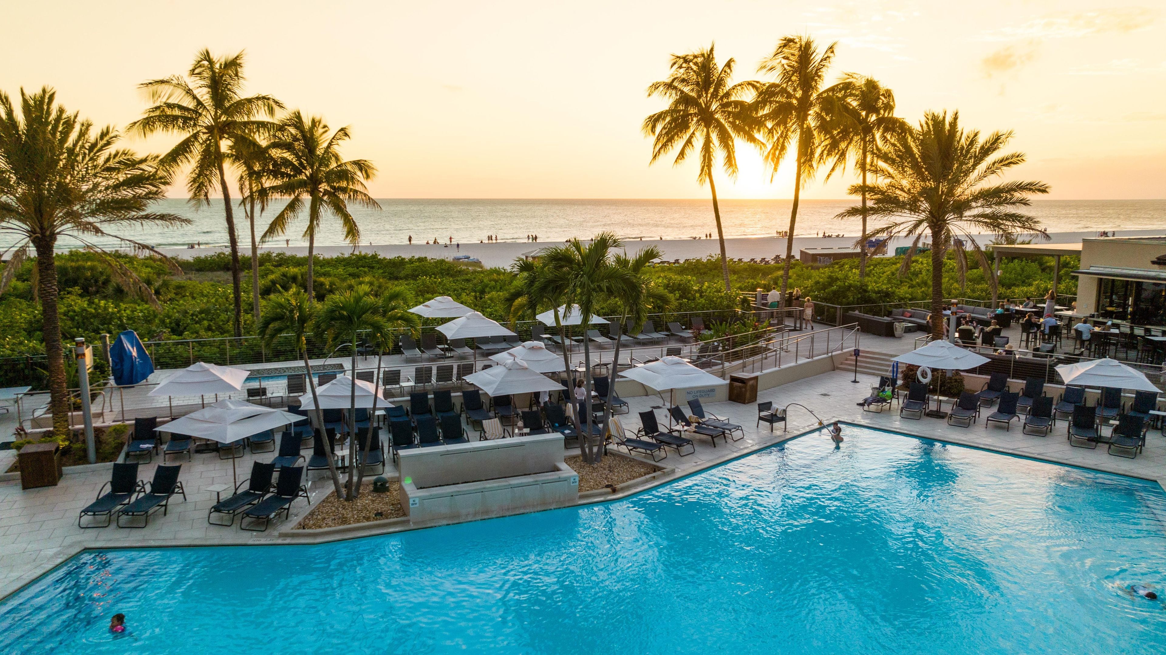 Ein Poolbereich mit Liegestühlen und Sonnenschirmen mit Blick auf einen Strand mit Palmen und dem Meer.