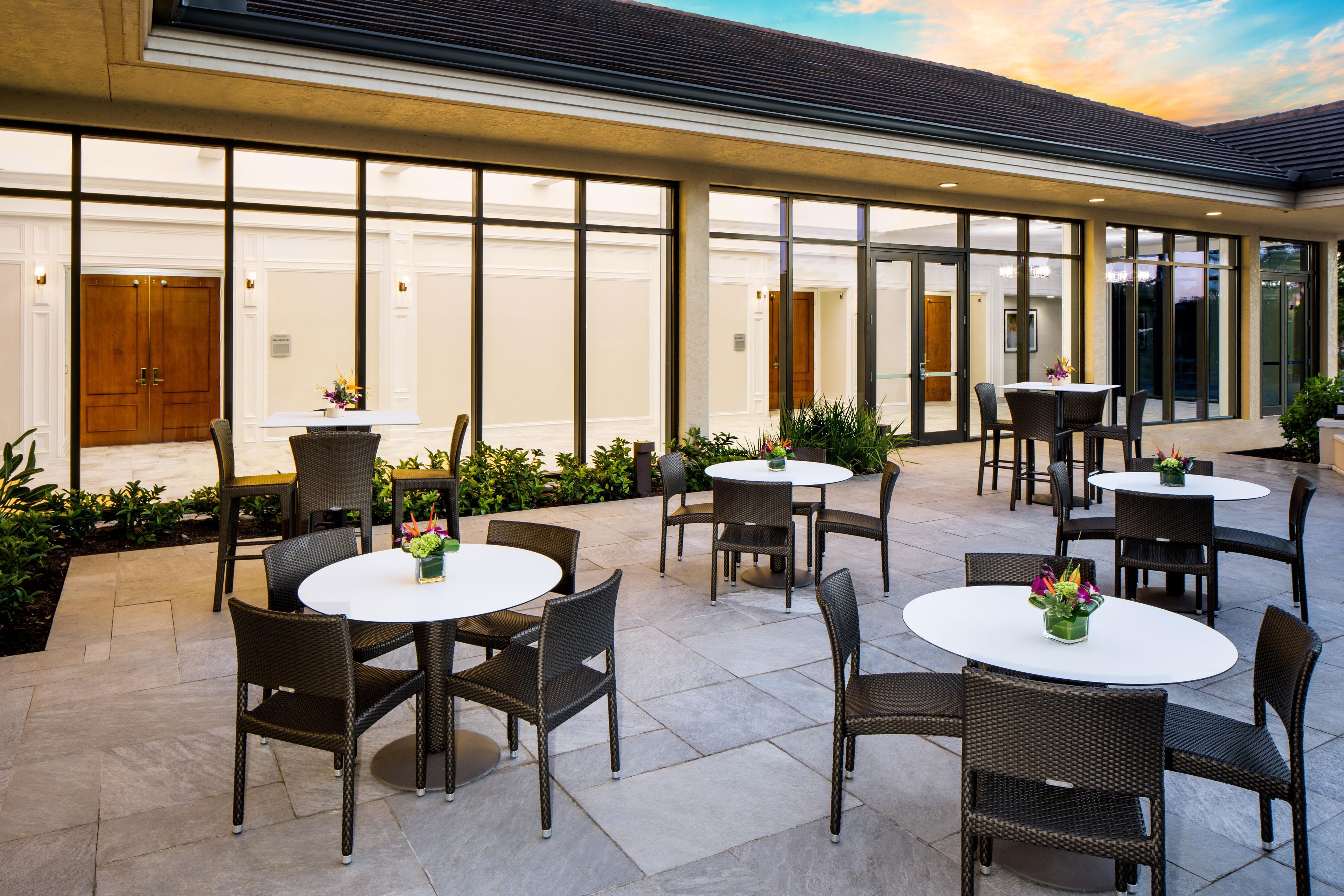 An outdoor patio area with round tables and wicker chairs, featuring a view of the building's interior through large glass doors.