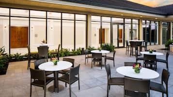 Un patio al aire libre con mesas redondas y sillas de mimbre, que ofrece vista del interior del edificio a través de amplios ventanales.